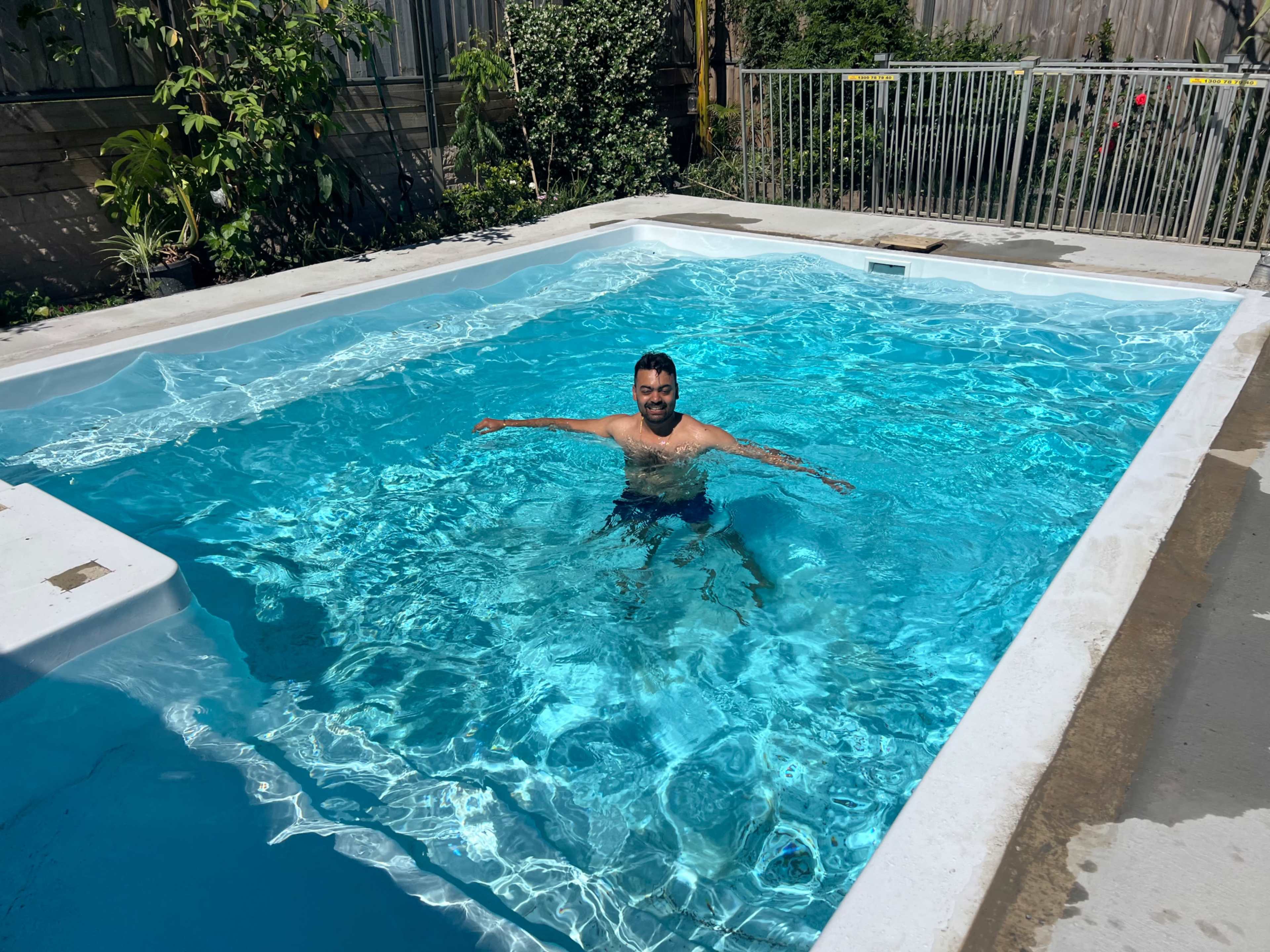 A man is swimming in a clear, blue pool surrounded by greenery and a fence.