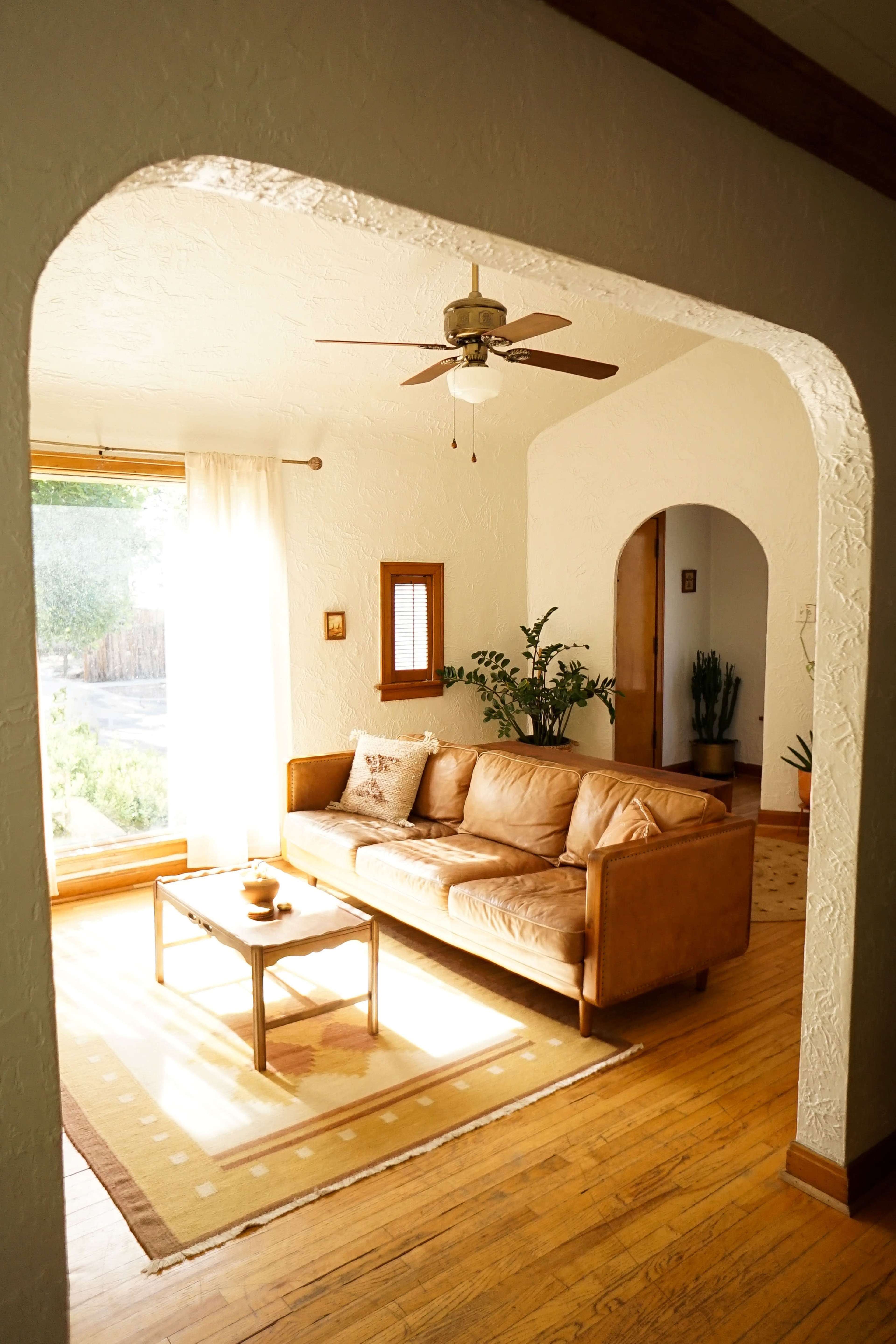 A spacious living room features a beige leather sofa, a wooden coffee table, and large windows allowing natural light to fill the space.