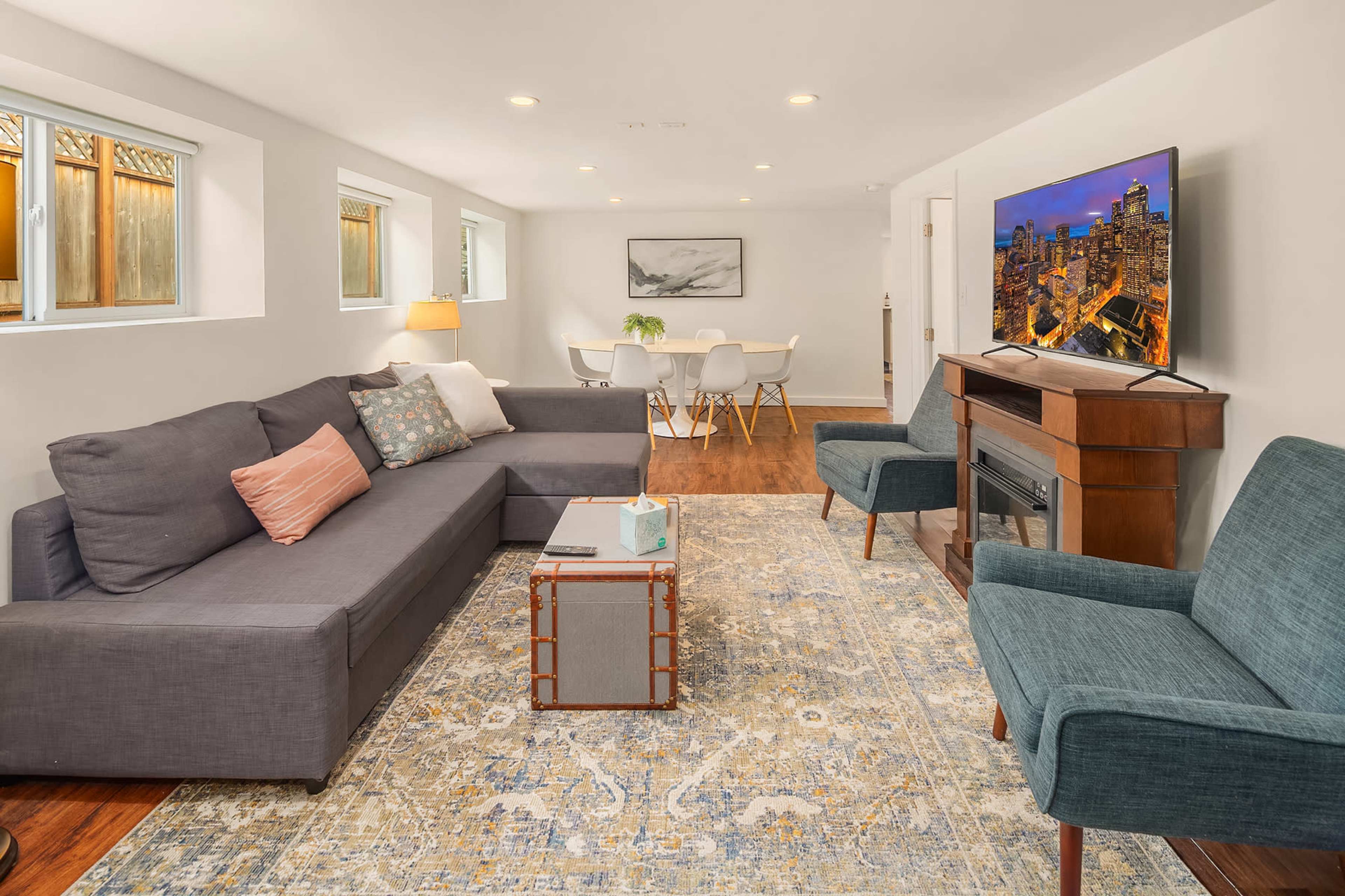 The image shows a modern living room with a sectional sofa, a coffee table, a flat-screen TV, and a dining area in the background.