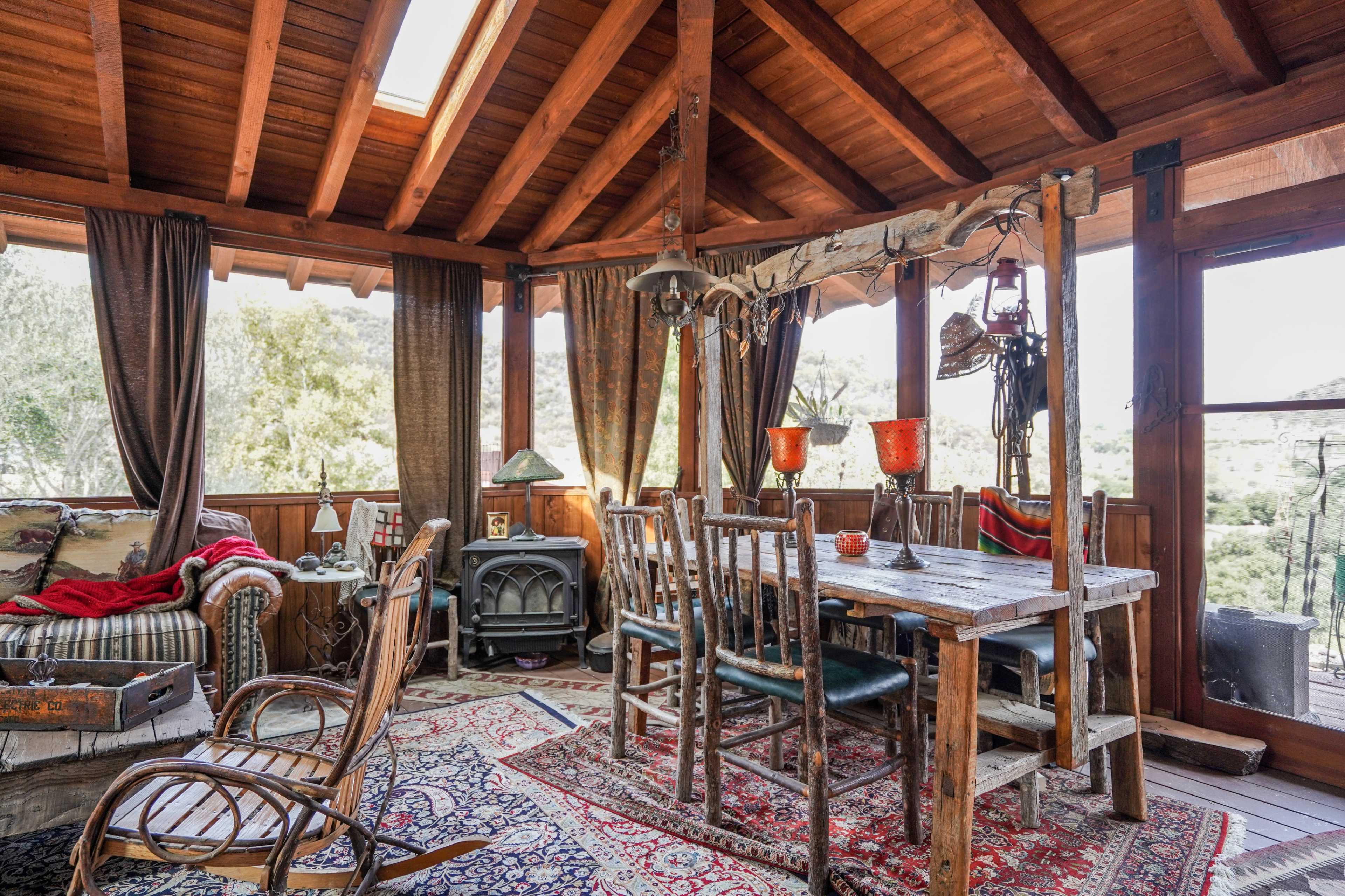The image shows a rustic dining area with wooden beams, a large table surrounded by chairs, and a cozy seating arrangement, all complemented by large windows revealing a natural landscape outside.