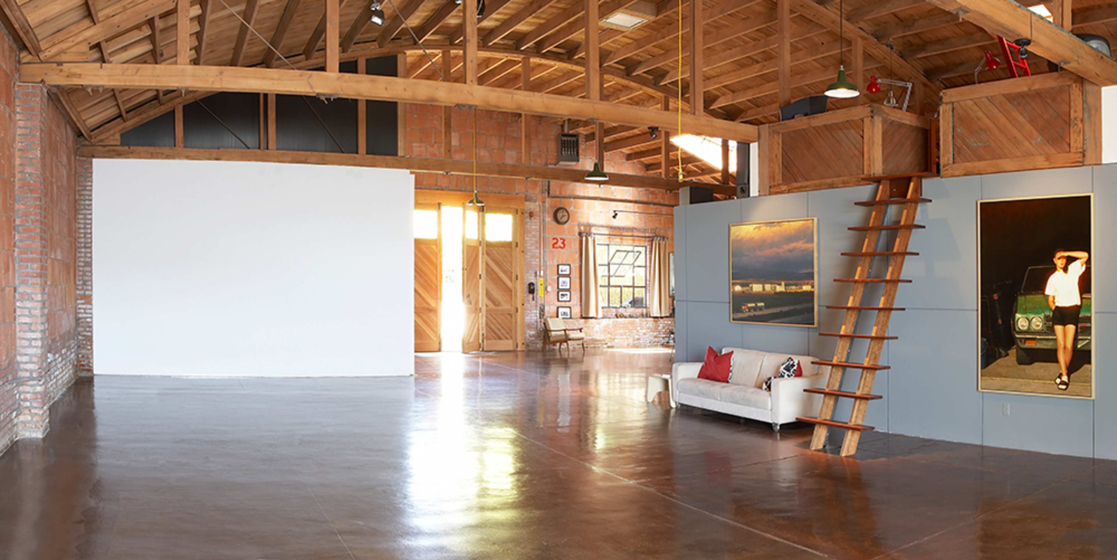 A spacious art gallery with a wooden ceiling, polished concrete floor, and an exhibition wall featuring a large photo alongside a staircase leading to a loft area.