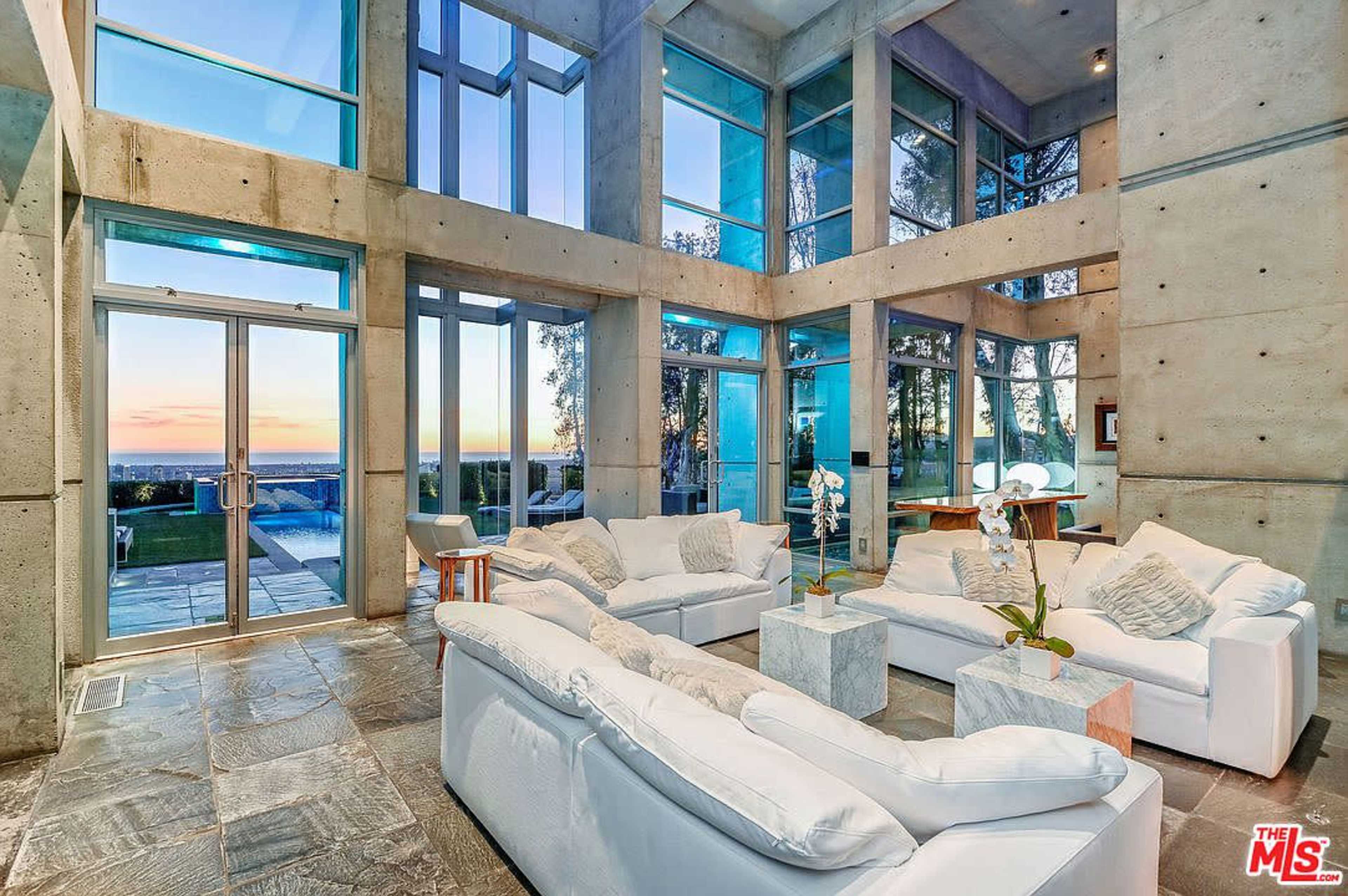 The image features a modern living room with large glass windows and concrete walls, showcasing a view of the outdoor landscape at sunset.