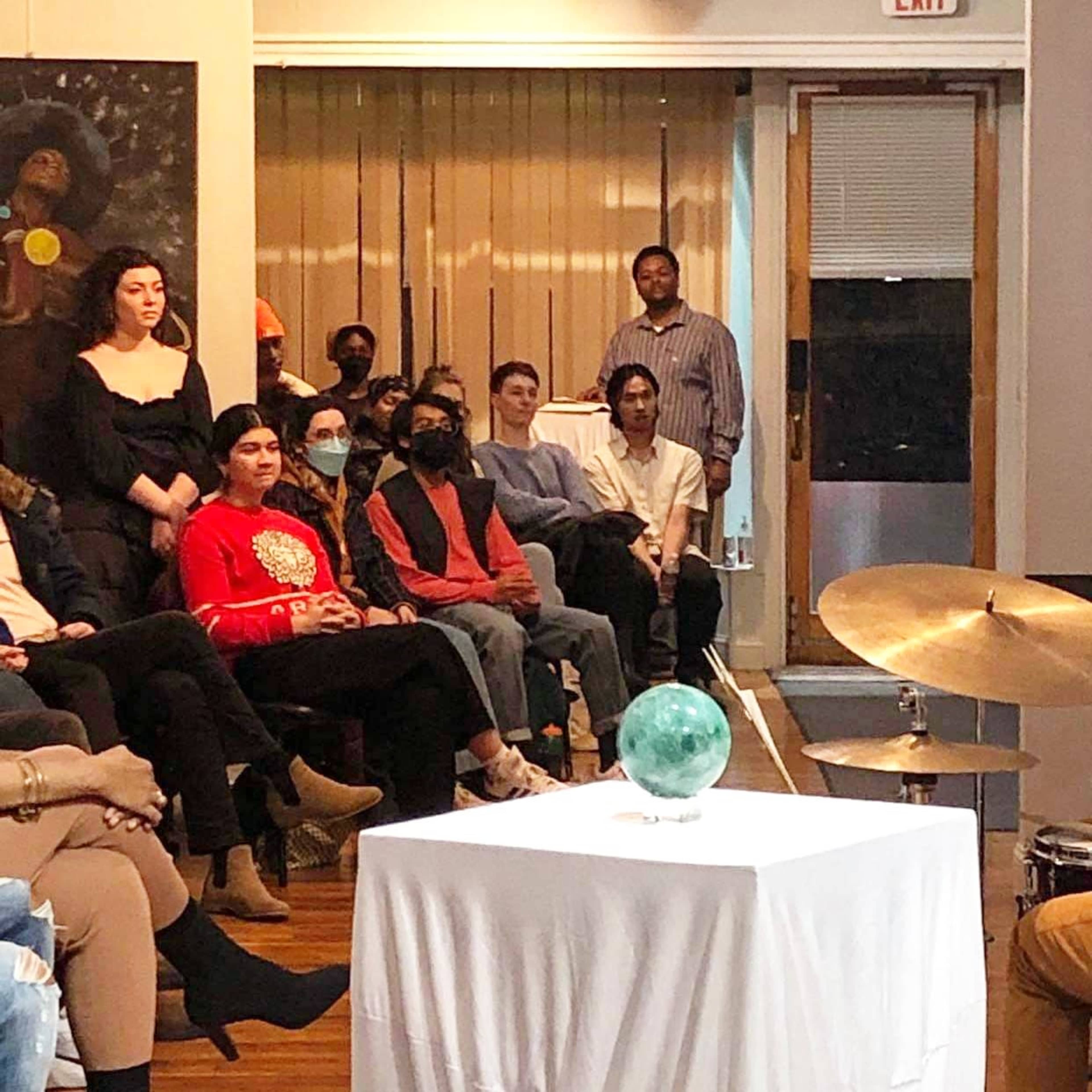 A group of people sits in a room facing a small table with a decorative glass orb, while a drummer is positioned nearby.