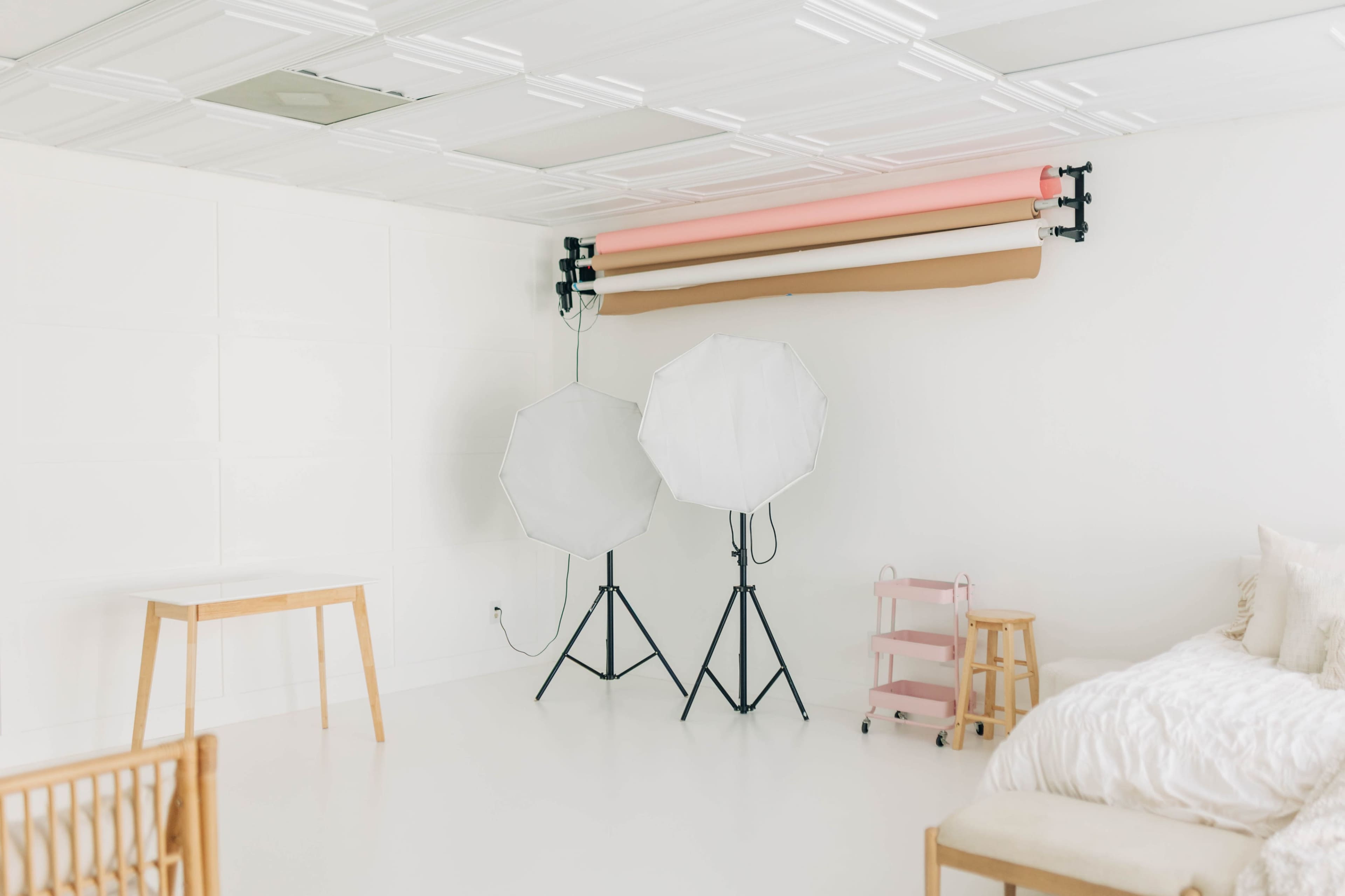 The image shows a bright, minimalist studio space equipped with two light stands, a rolling cart, a stool, and backdrops mounted on a wall.