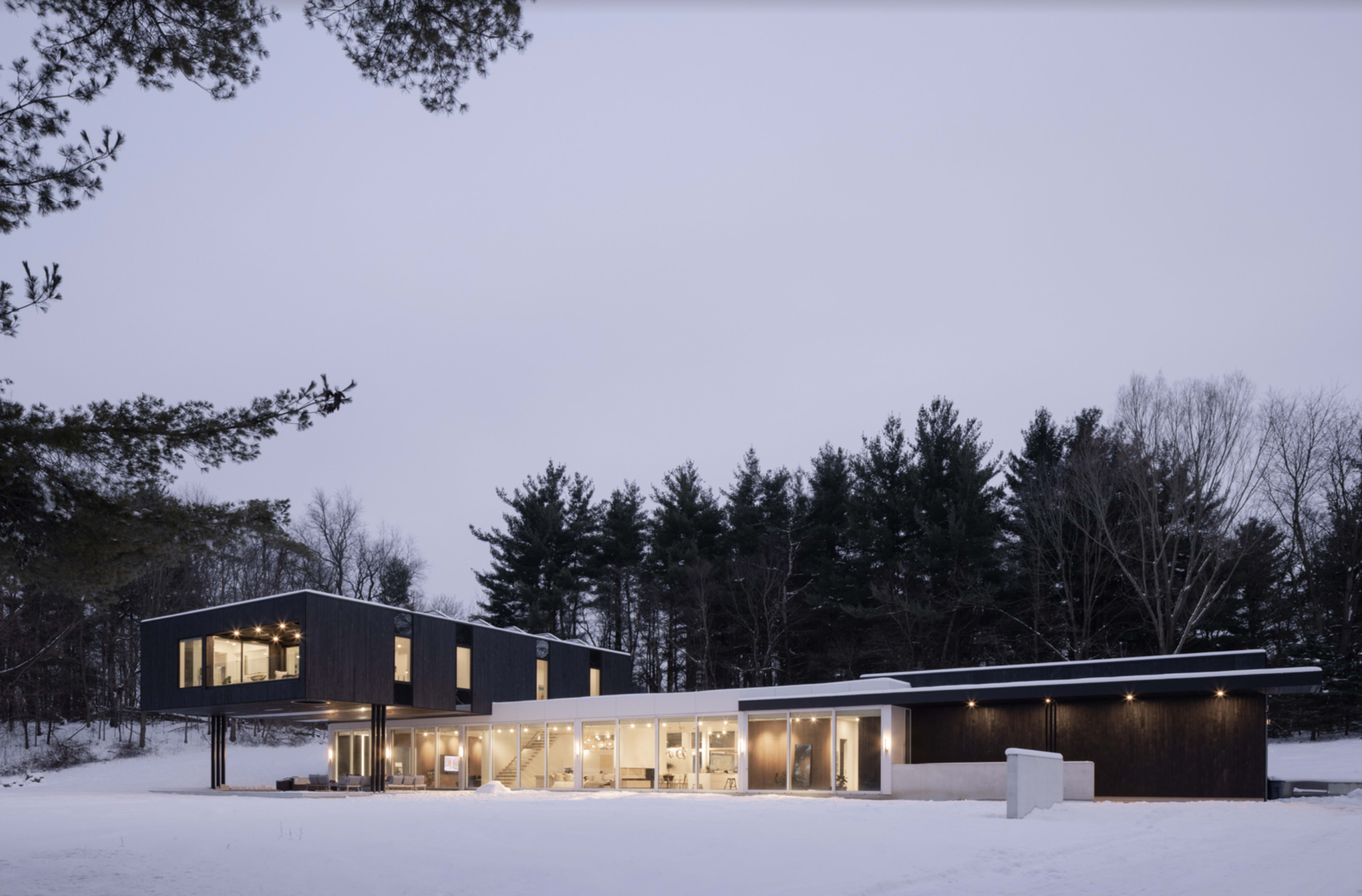 A modern, black exterior house sits in a snowy landscape surrounded by trees.