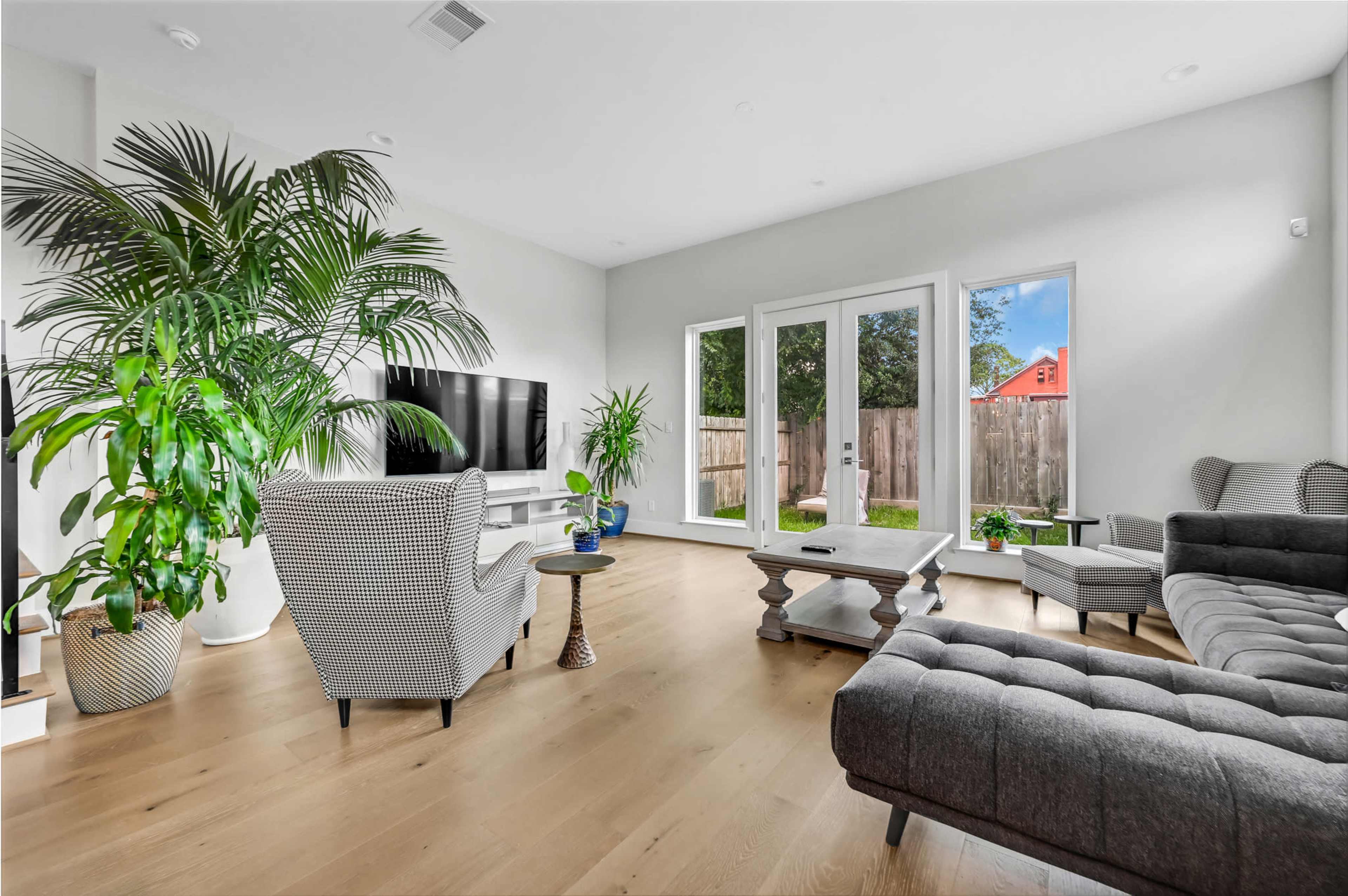 The living room features a large television, modern furniture, and several potted plants by large windows that overlook a fenced backyard.