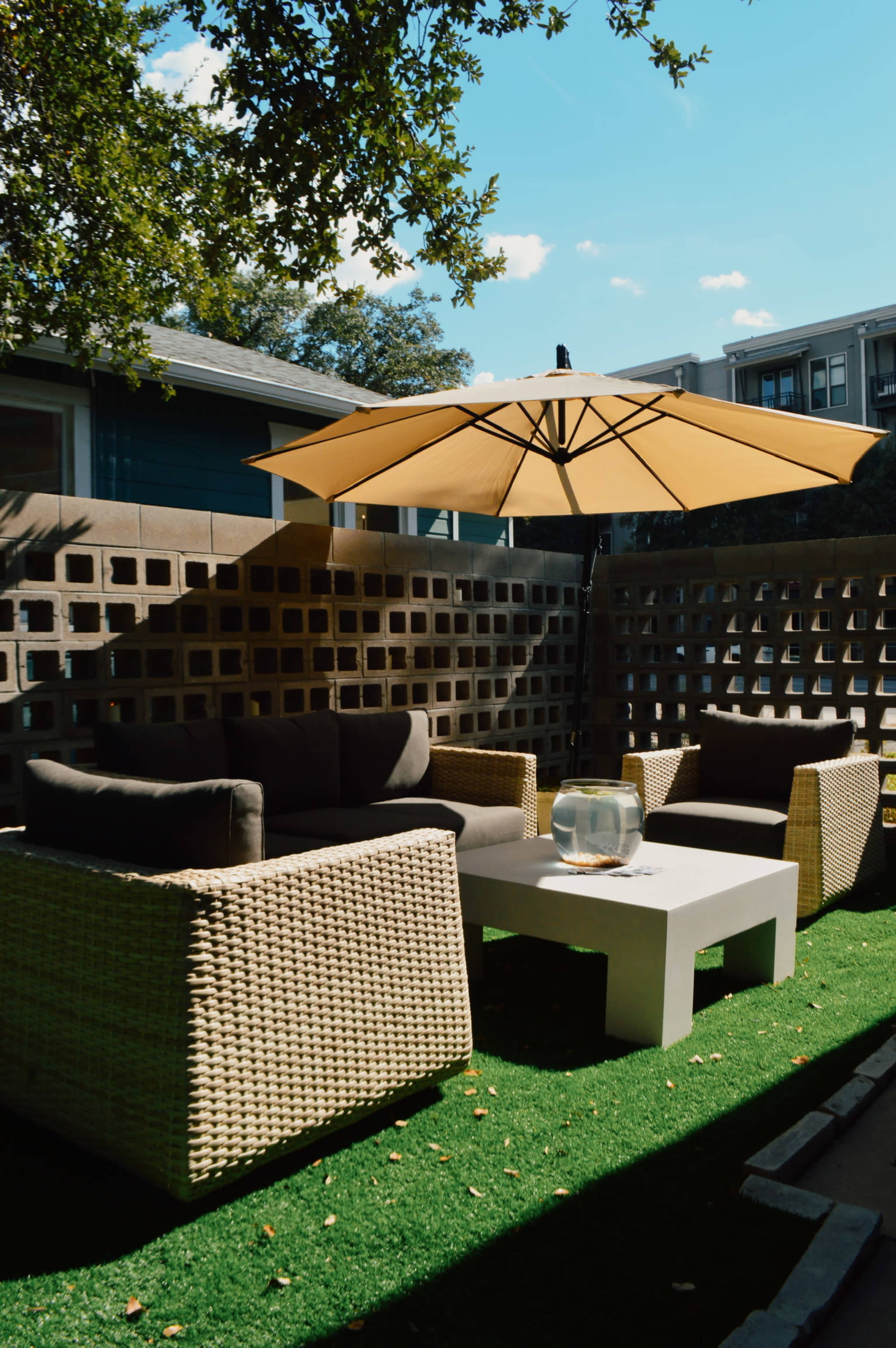 A patio area features two wicker chairs and a white table beneath a large yellow umbrella, surrounded by a decorative stone wall and artificial grass.