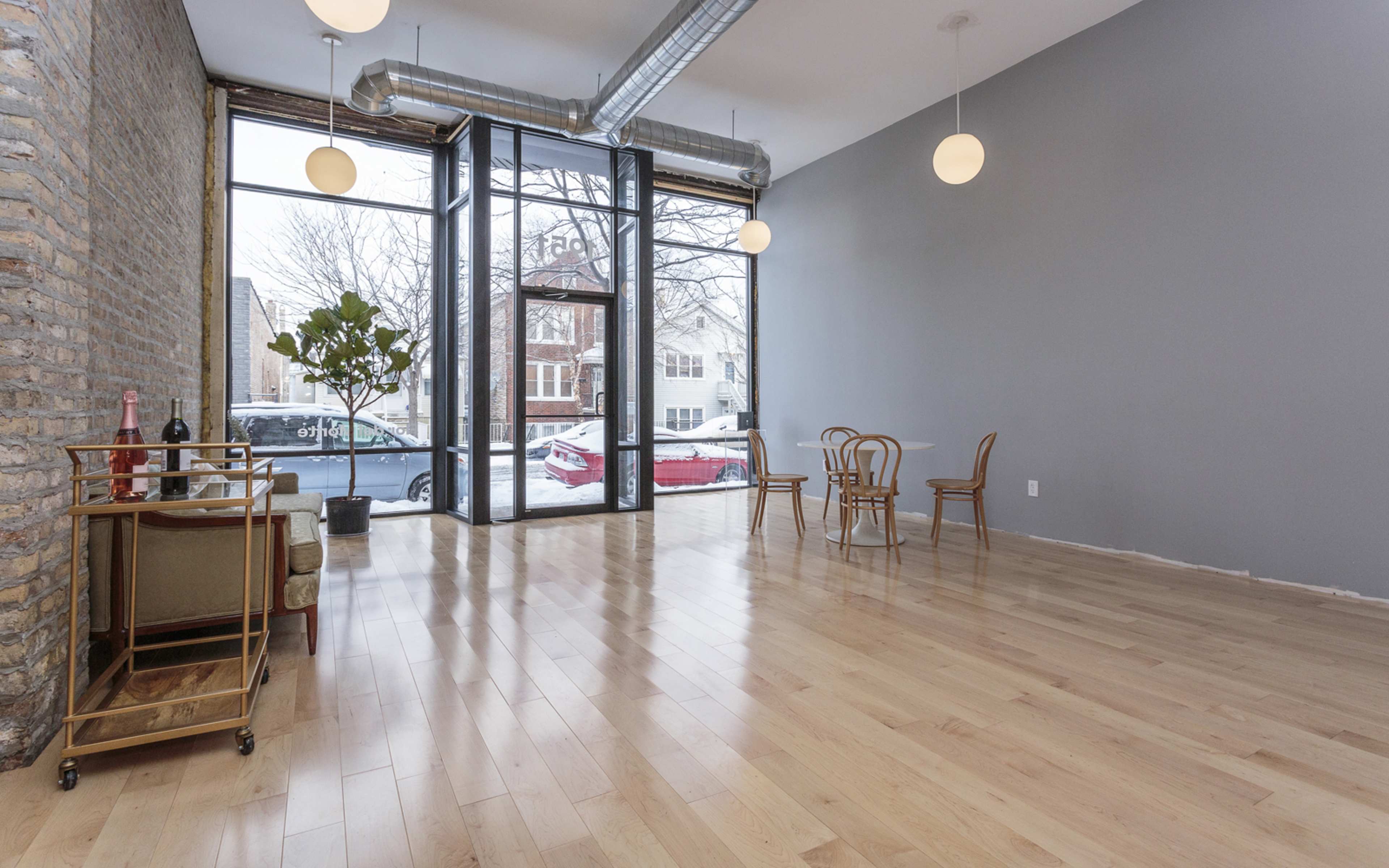 An empty room with large windows, a polished wooden floor, and a small dining table with chairs.