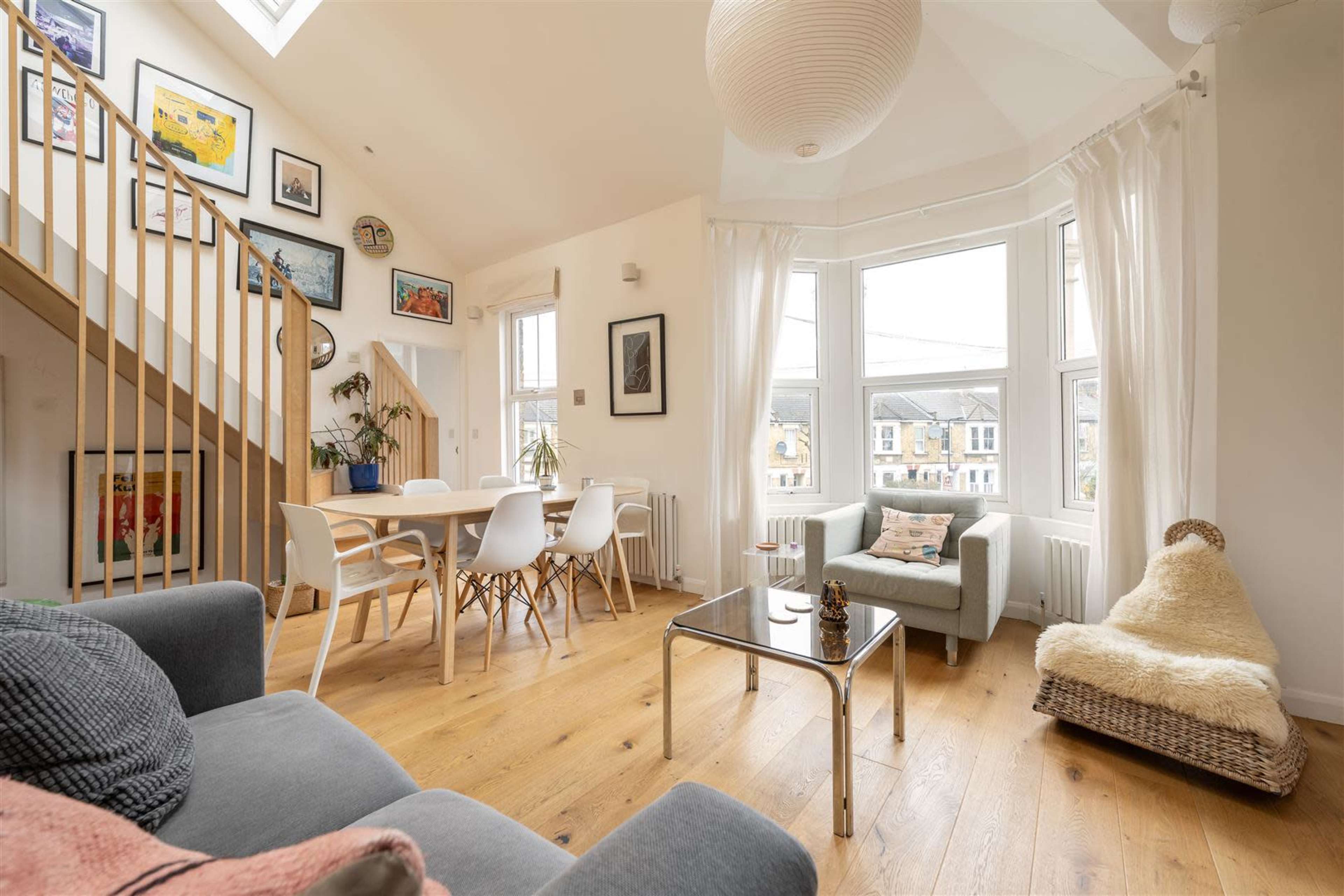 The image shows a bright, open living space featuring a staircase, dining area, and seating arrangements with large windows allowing natural light to fill the room.