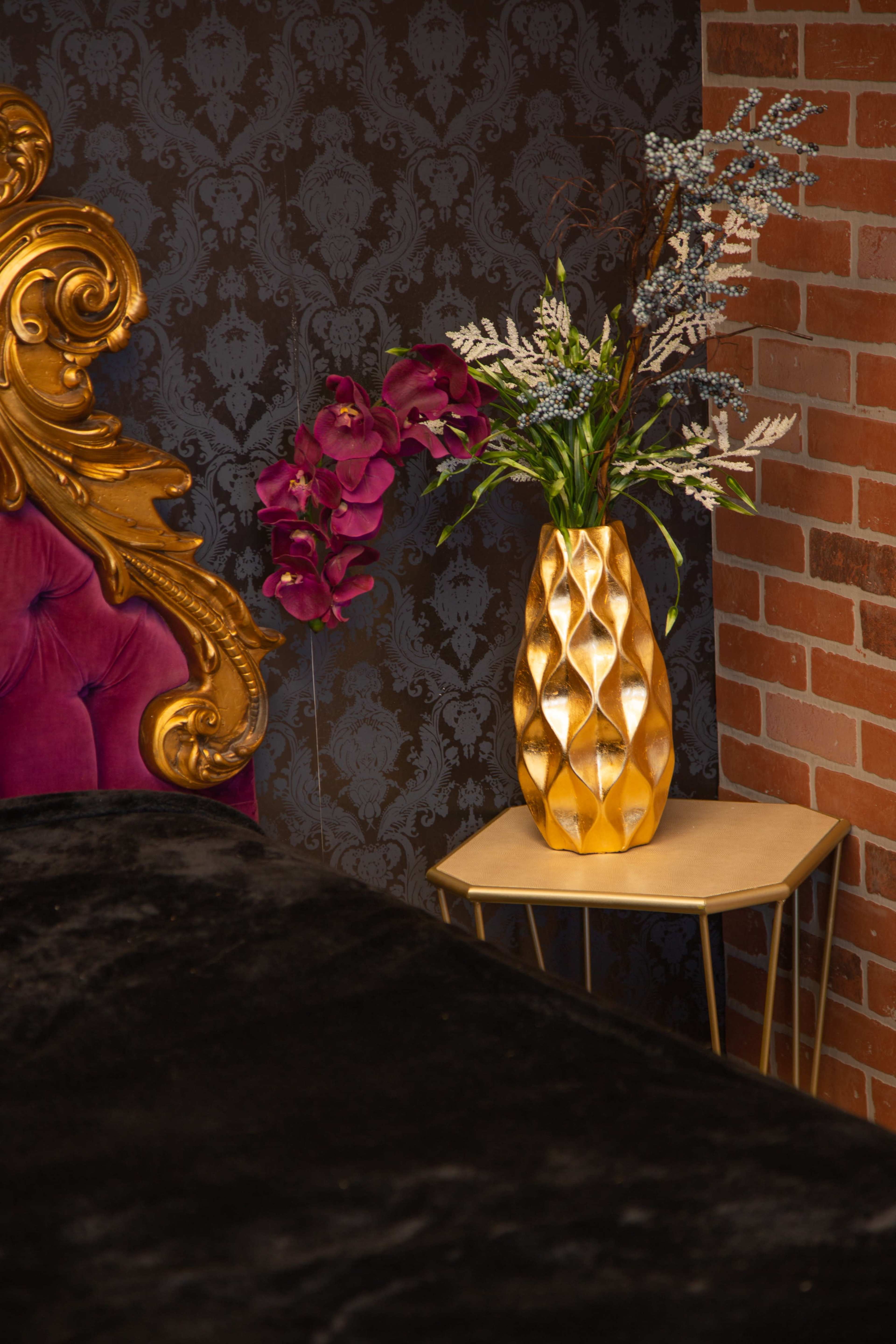 A gold vase filled with flowers sits on a small table next to an ornate, purple-upholstered bed against a patterned wallpaper and brick wall backdrop.