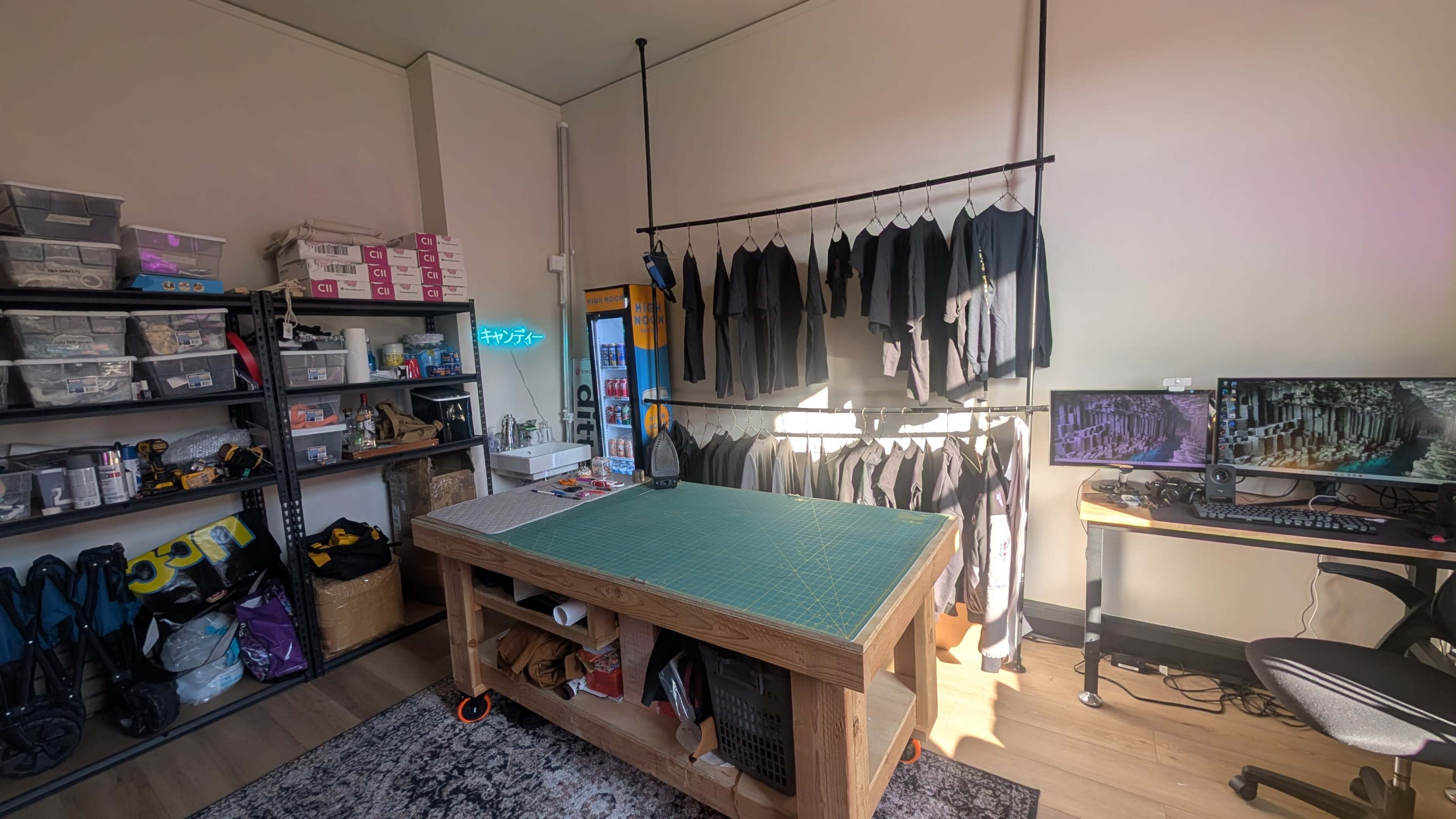 The image shows a well-organized workspace with a large table, hanging clothing items, shelves with storage boxes, and a computer desk set up in a brightly lit room.
