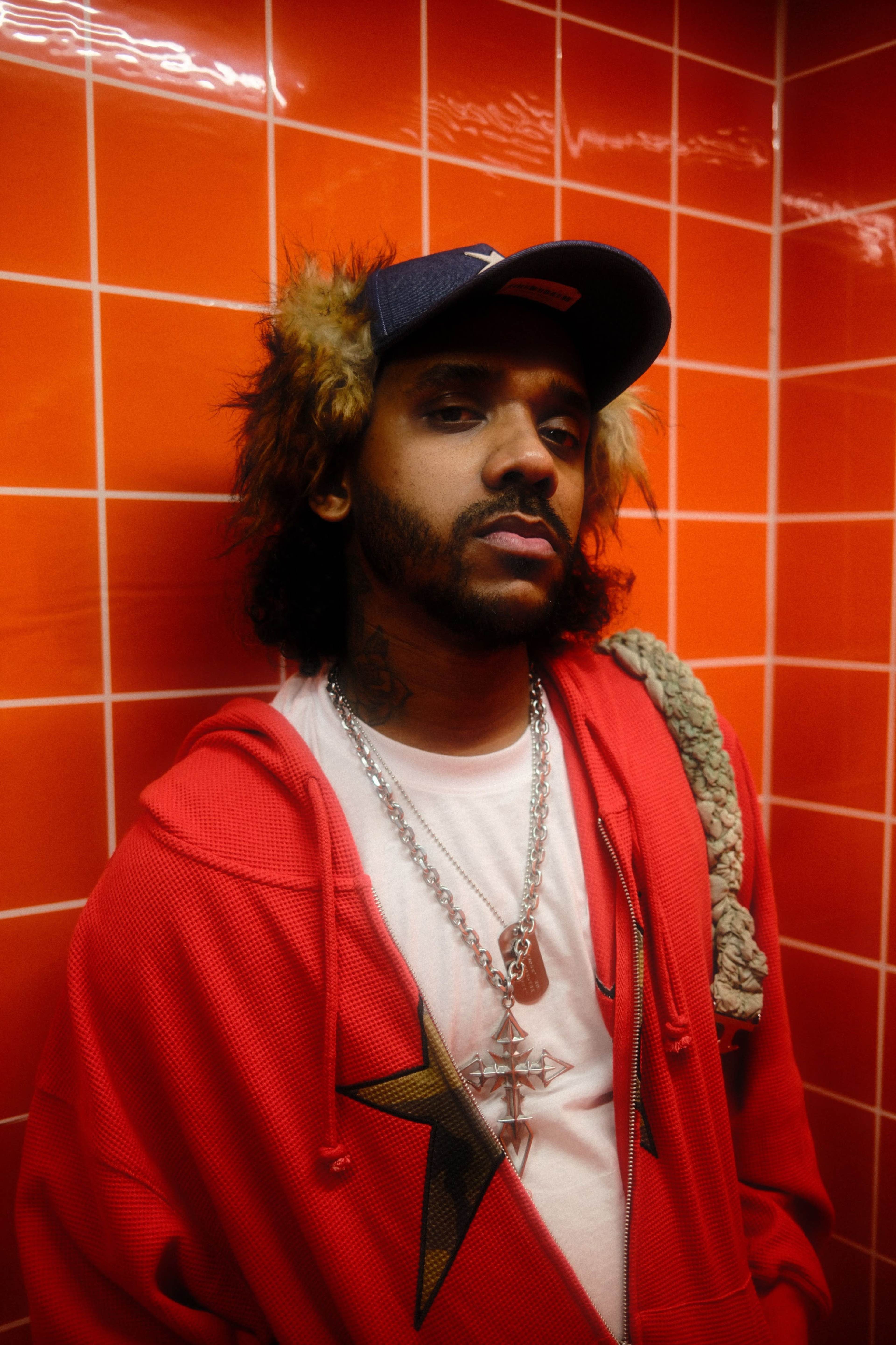 A person wearing a red hoodie and a blue cap stands against a bright orange tiled wall.