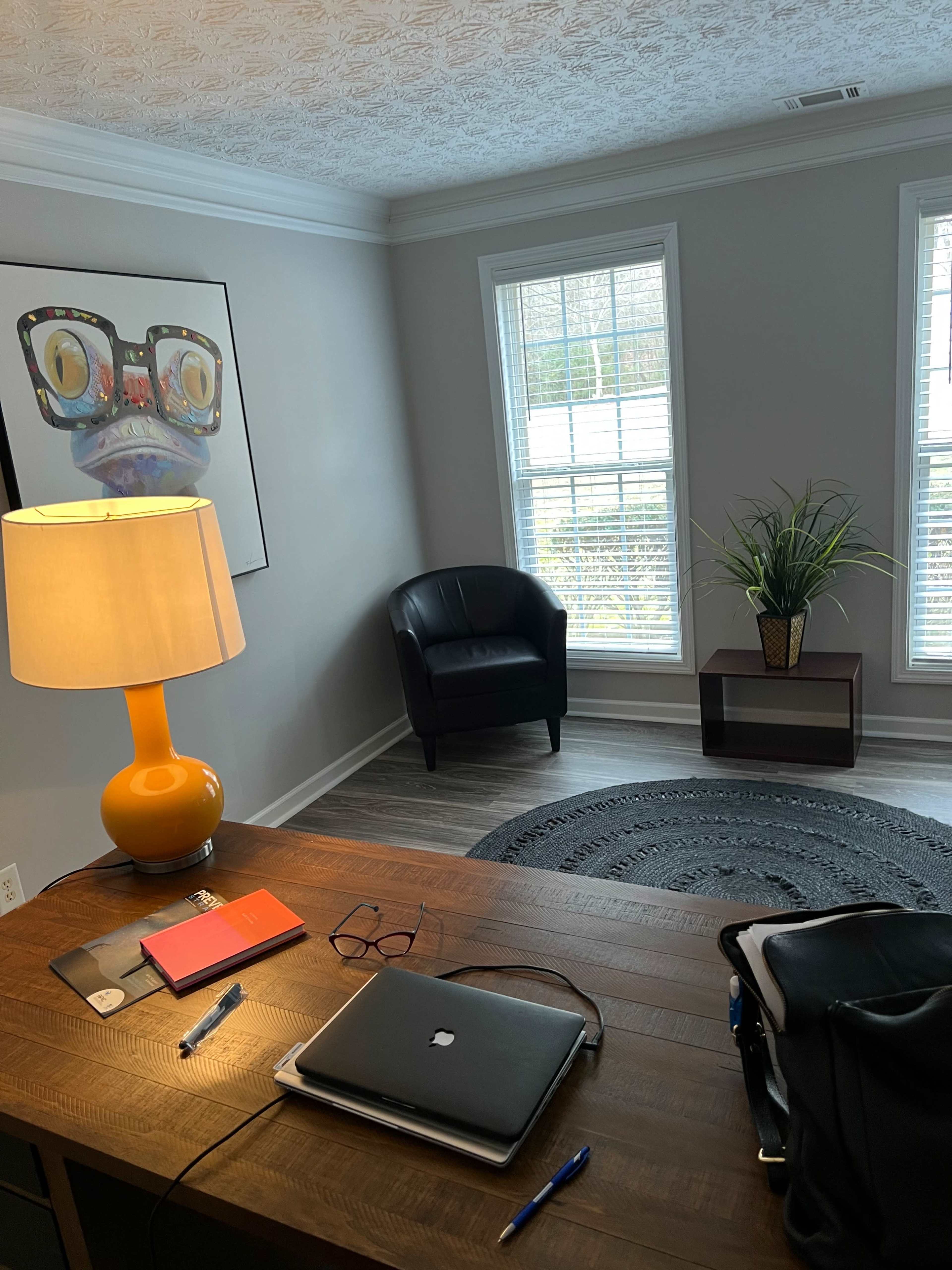 The image shows a modern home office with a wooden desk, a laptop, and a lamp, alongside a chair and a plant near the windows.