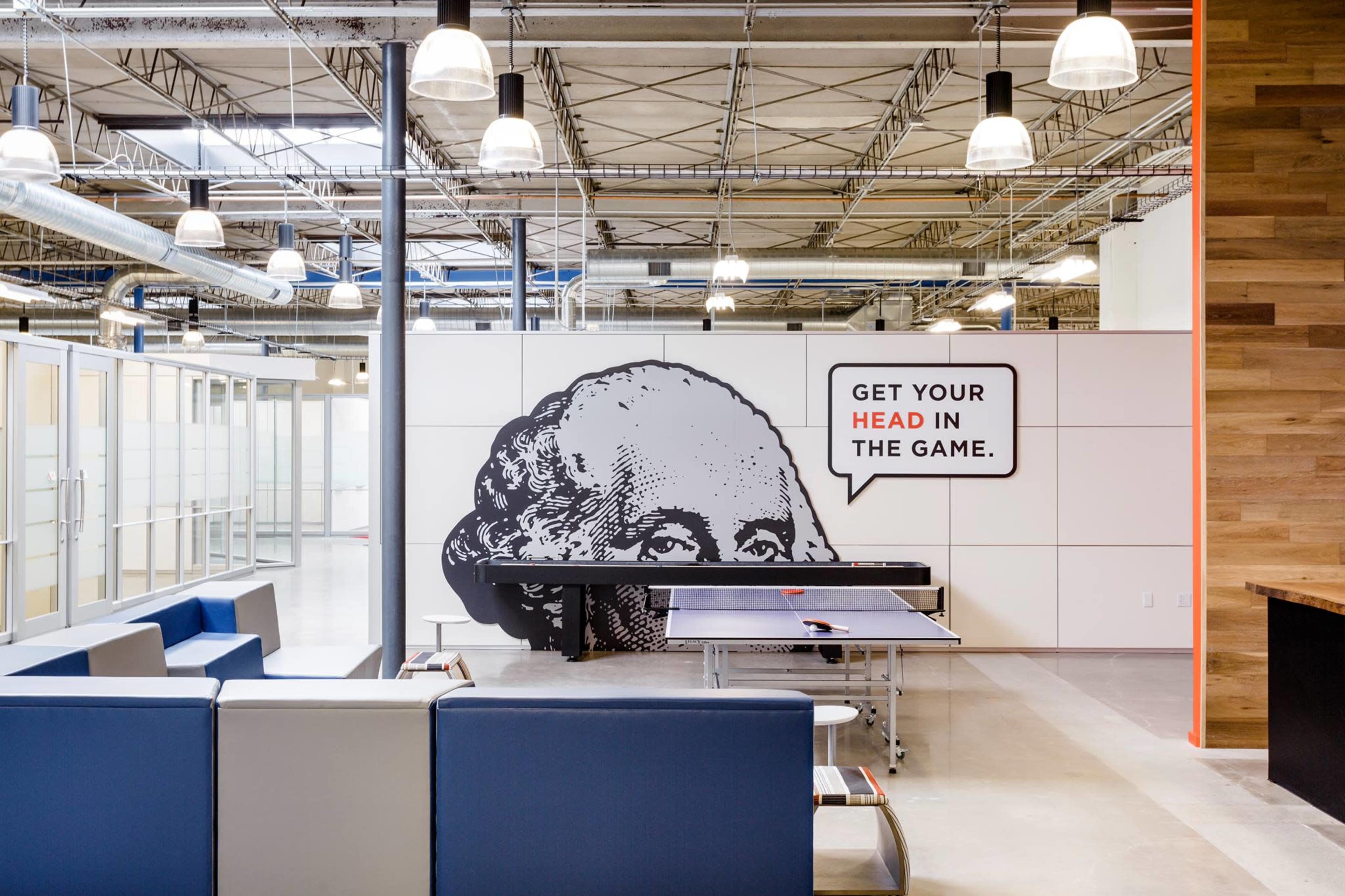 A modern office space featuring a ping pong table, lounge seating, and a wall graphic of George Washington with the phrase "GET YOUR HEAD IN THE GAME."