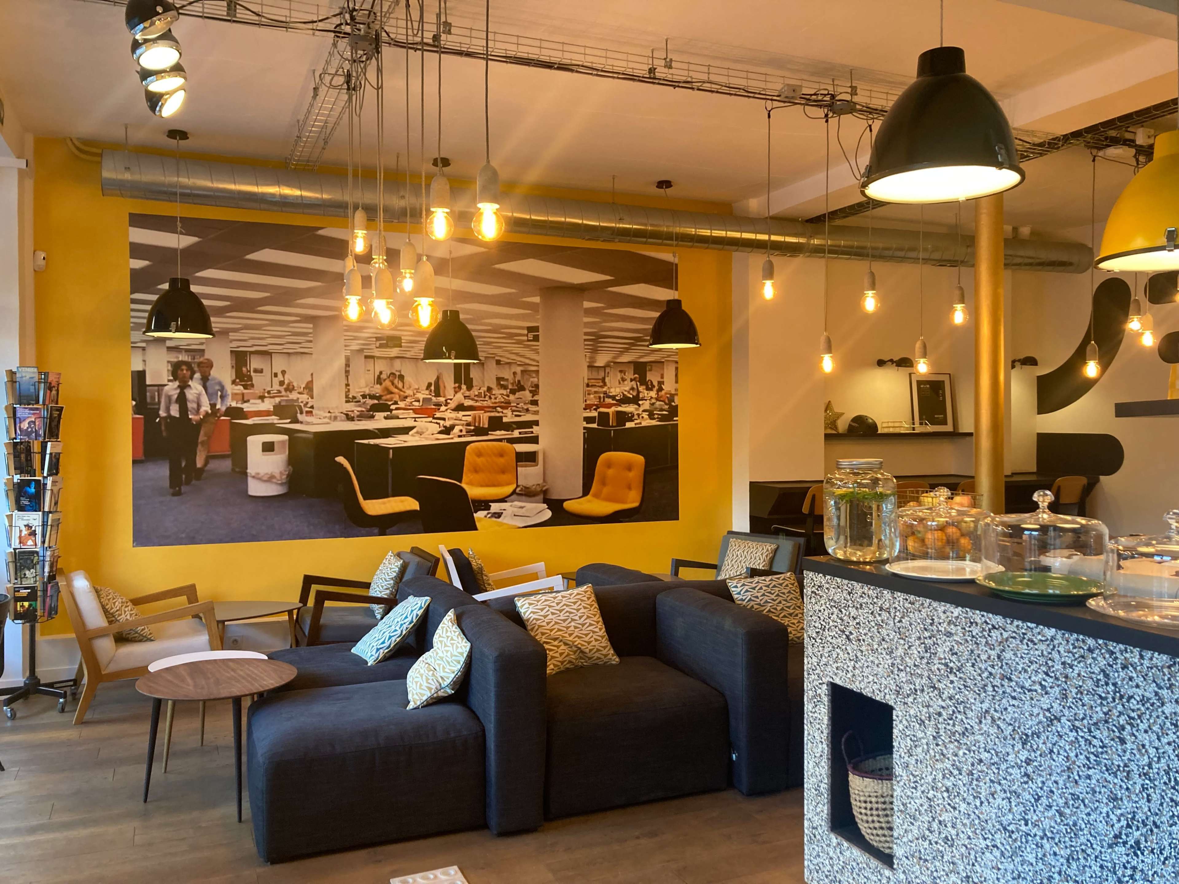 The image shows a modern lounge area with a mix of seating, bright yellow walls, and a large mural of an office environment.