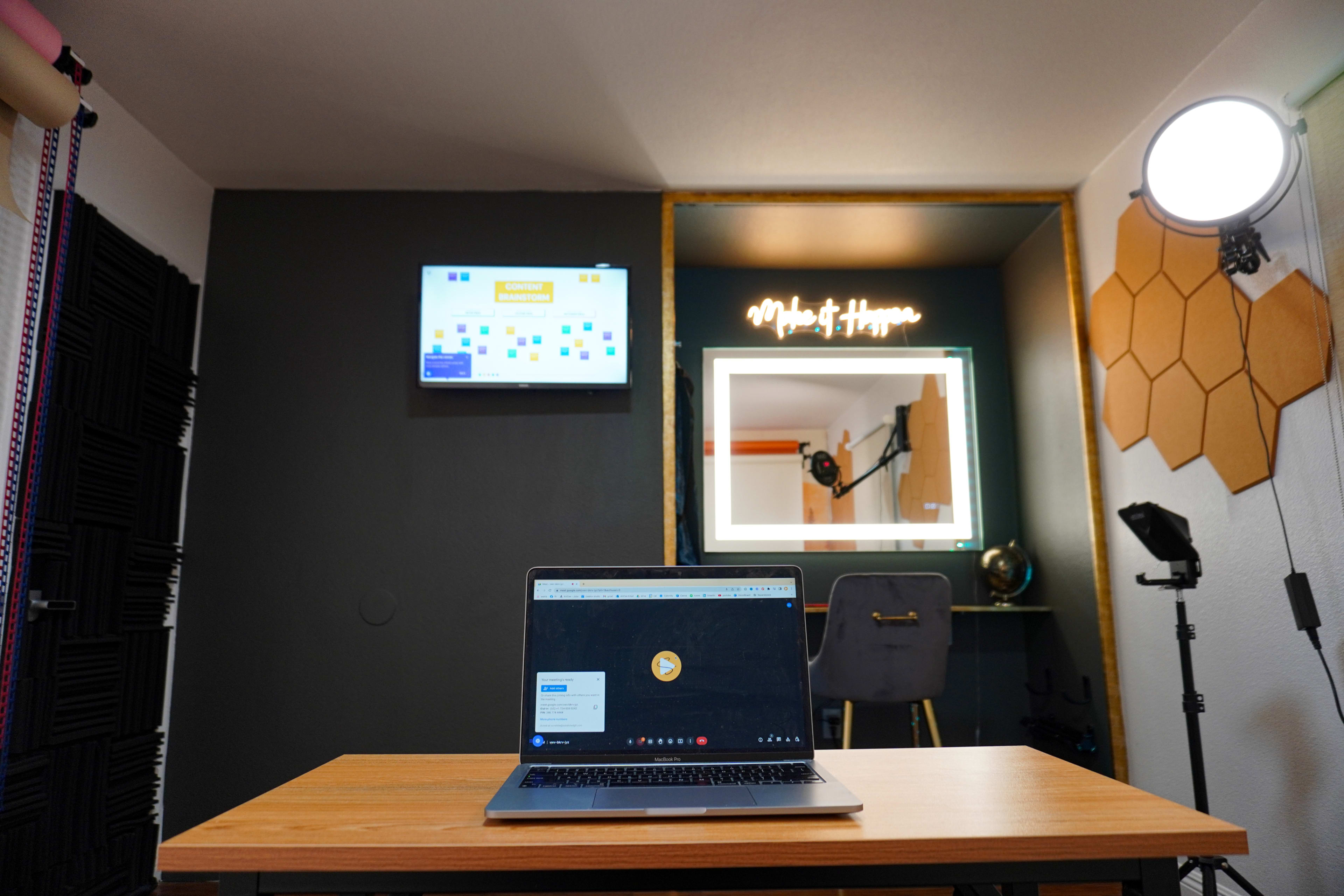 A laptop sits on a wooden table in a room featuring a mirror, a monitor on the wall, and various studio equipment.