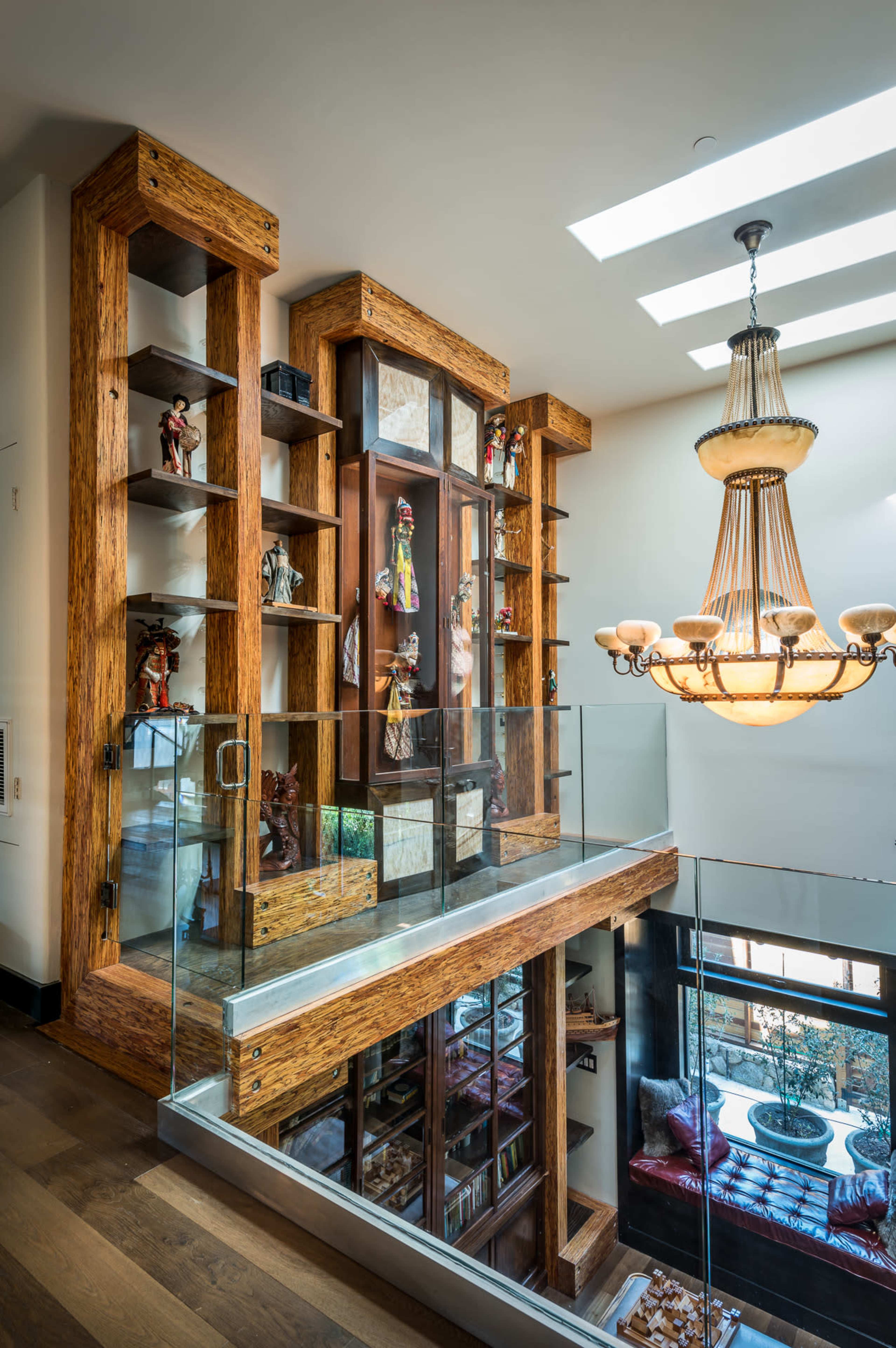 A modern interior with a glass railing, wooden shelves housing figurines, and a decorative chandelier above.