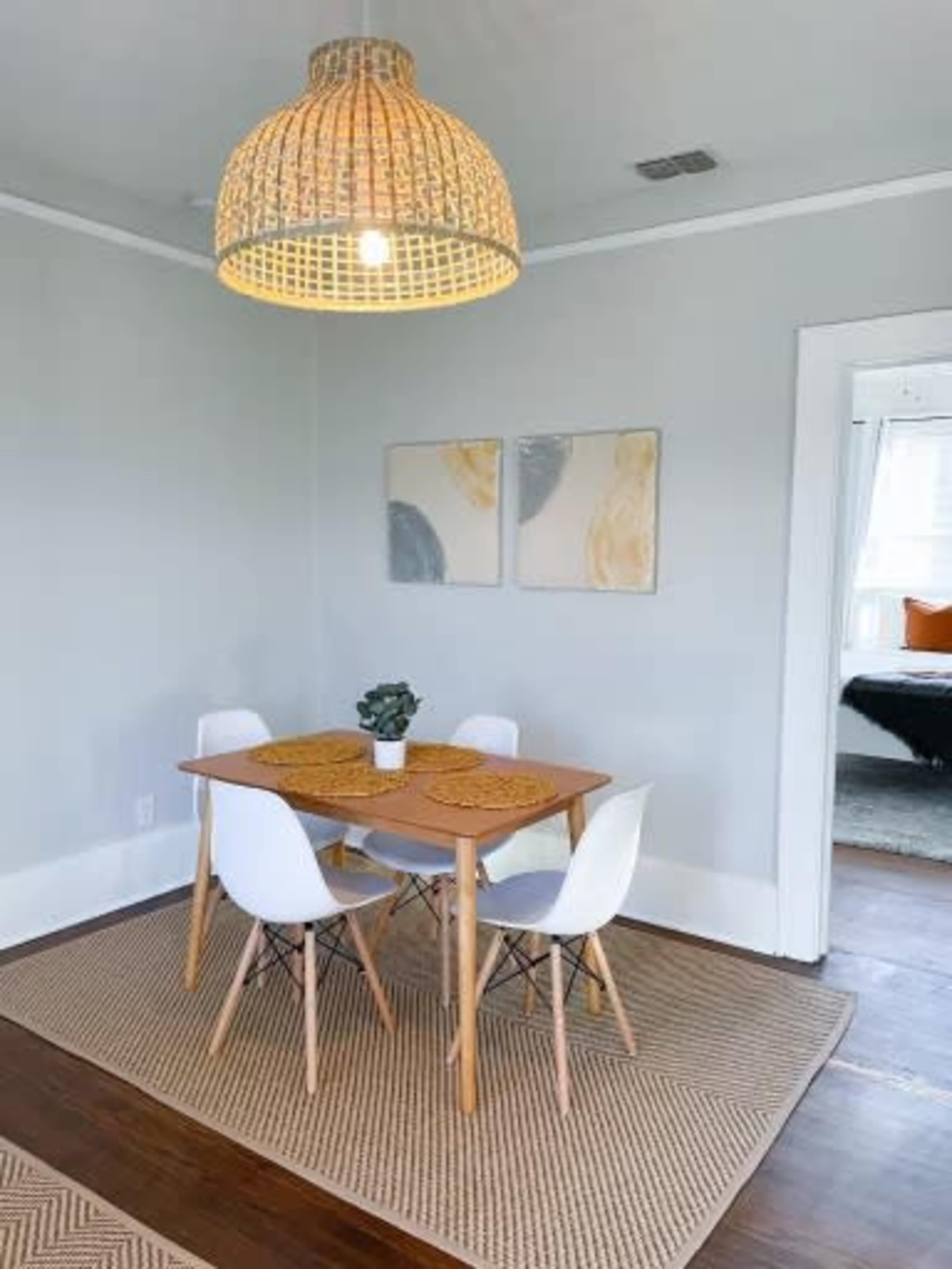 A small dining area features a wooden table surrounded by four white chairs, with a pendant light overhead and two framed artworks on the wall.
