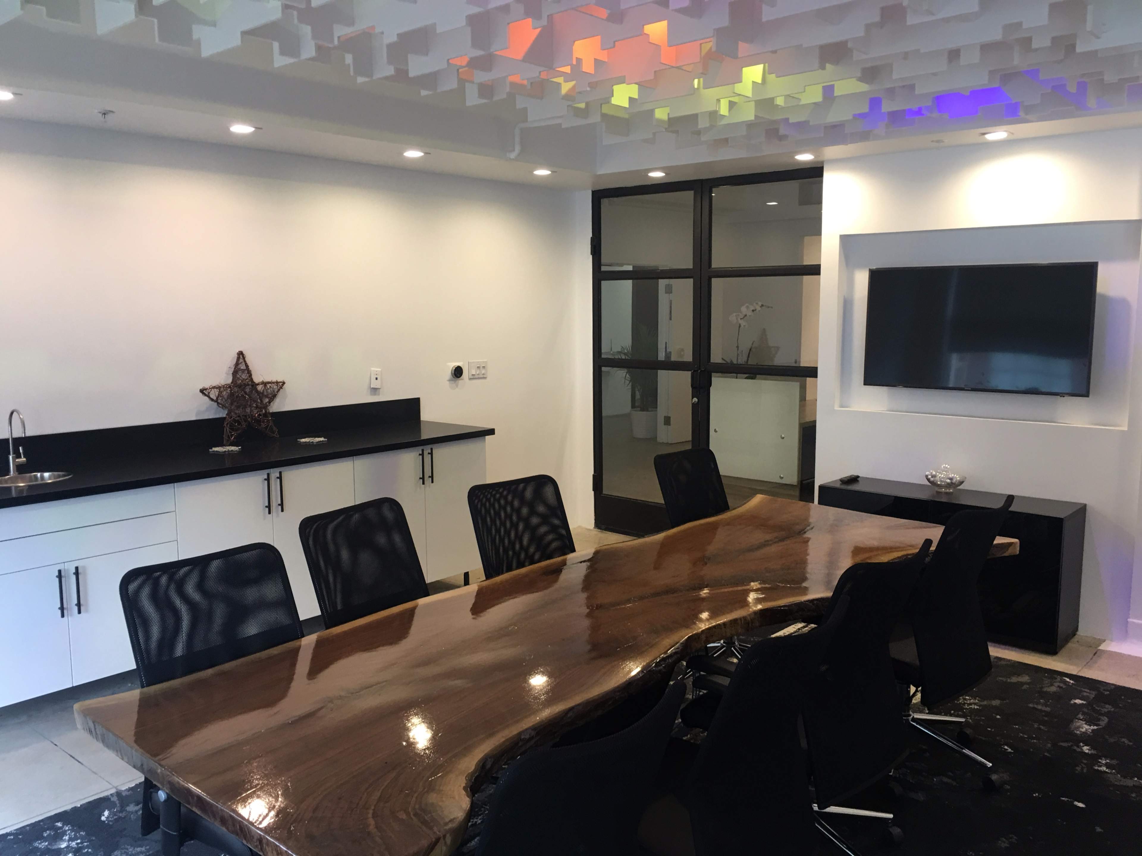 A modern meeting room features a large wooden conference table surrounded by black chairs, with a television and a kitchen area in the background.