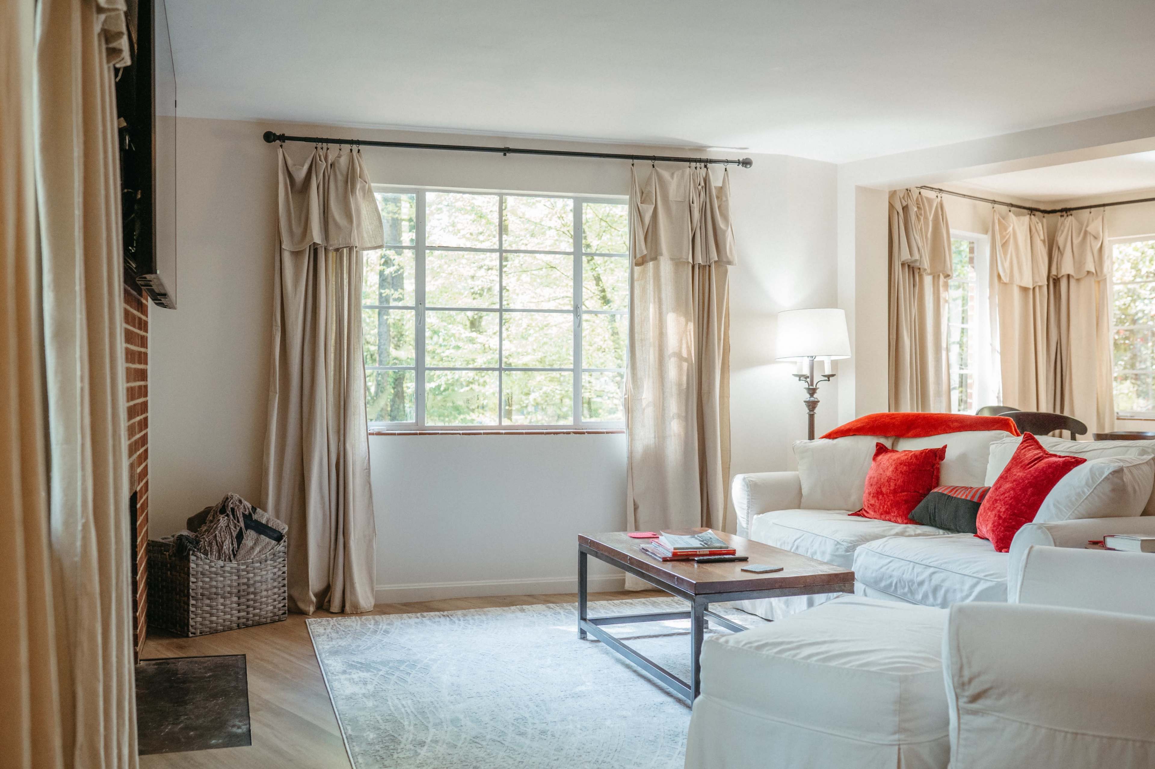A well-lit living room features a white sofa with red accents, a wooden coffee table, and large windows draped with soft curtains.