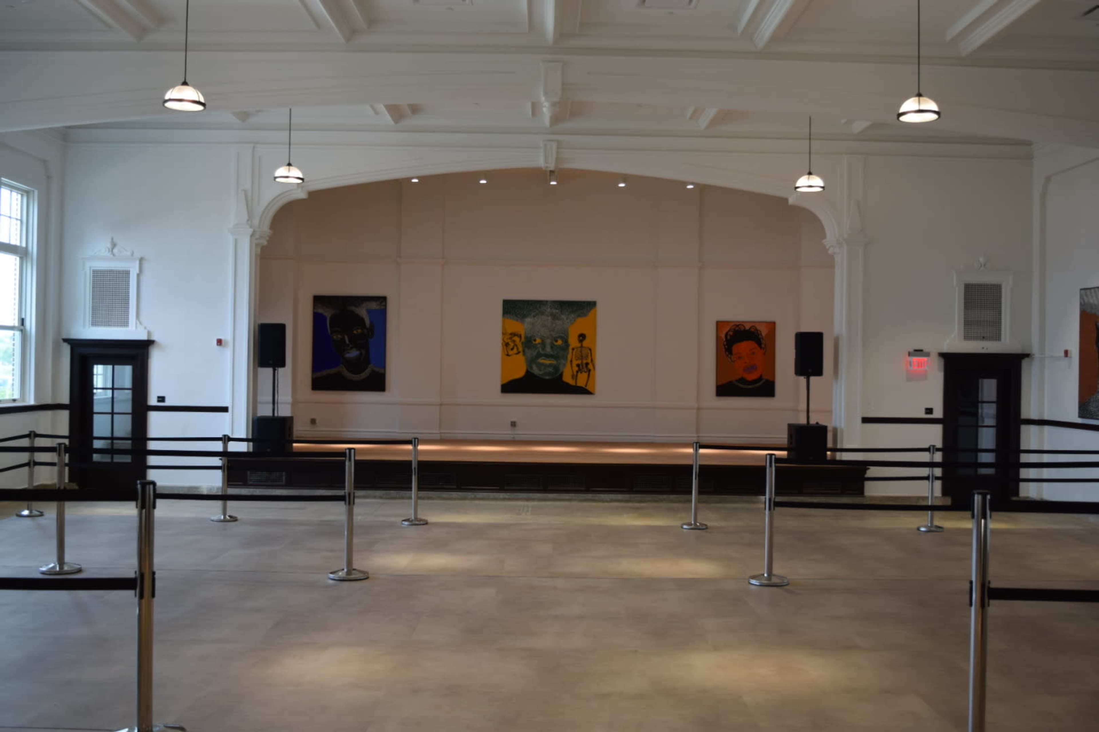 The image shows a spacious, empty hall with an elevated stage and paintings displayed on the wall.