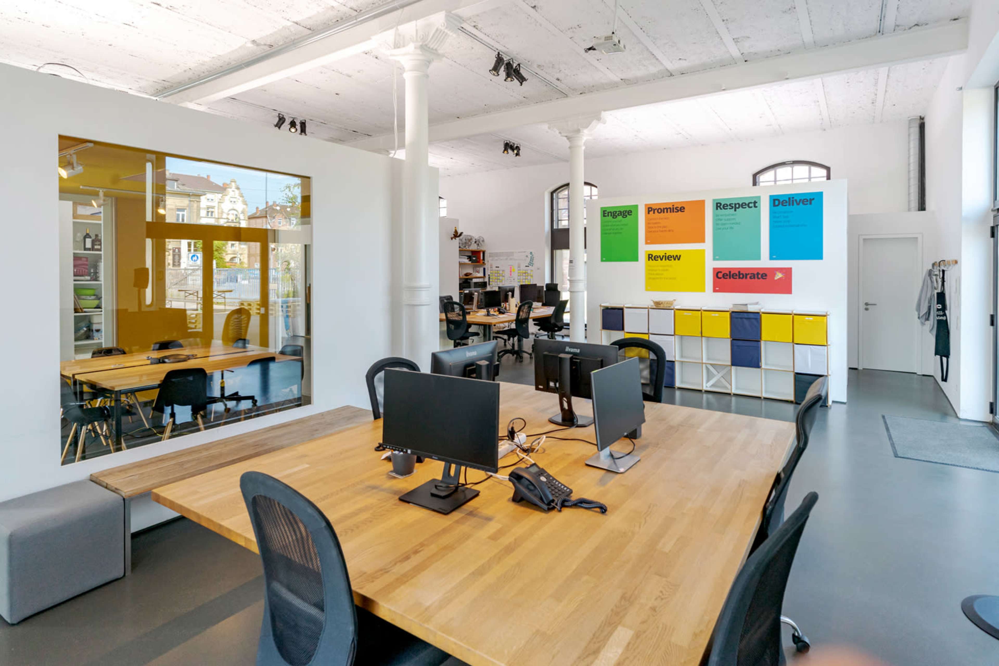 A modern office space features a large wooden table with multiple computer workstations and colorful wall displays promoting company values.