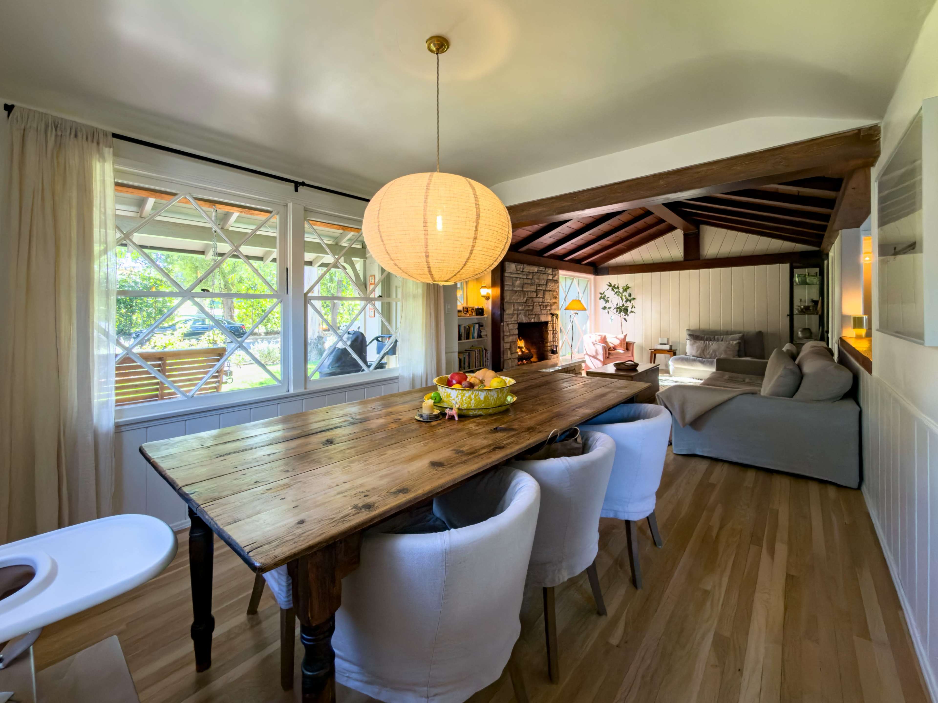 A wooden dining table with round chairs is set in a bright room that features large windows and a cozy seating area.