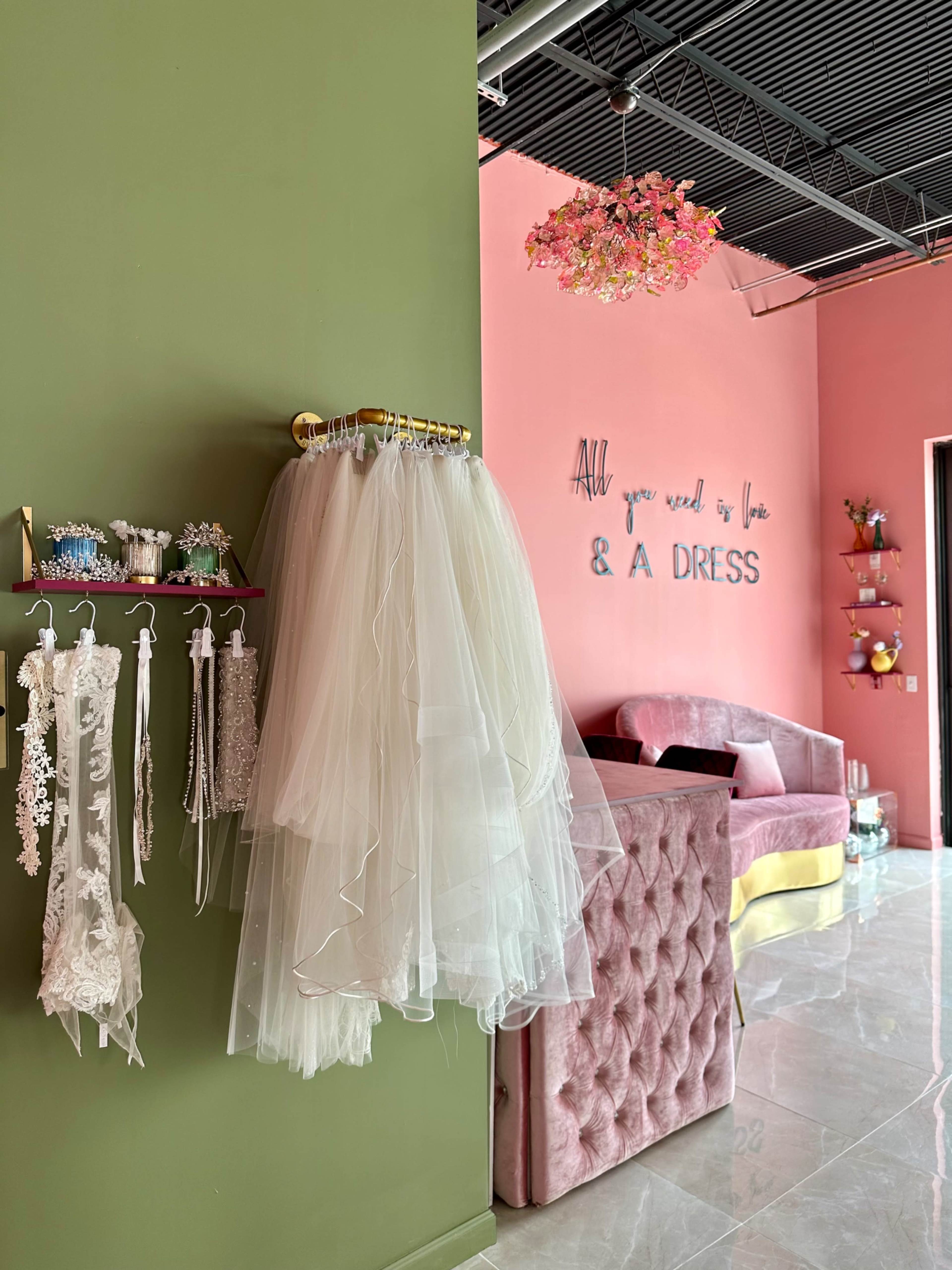 A boutique interior features a green wall with hanging bridal accessories and a pink wall adorned with a decorative phrase, alongside a plush seating area.
