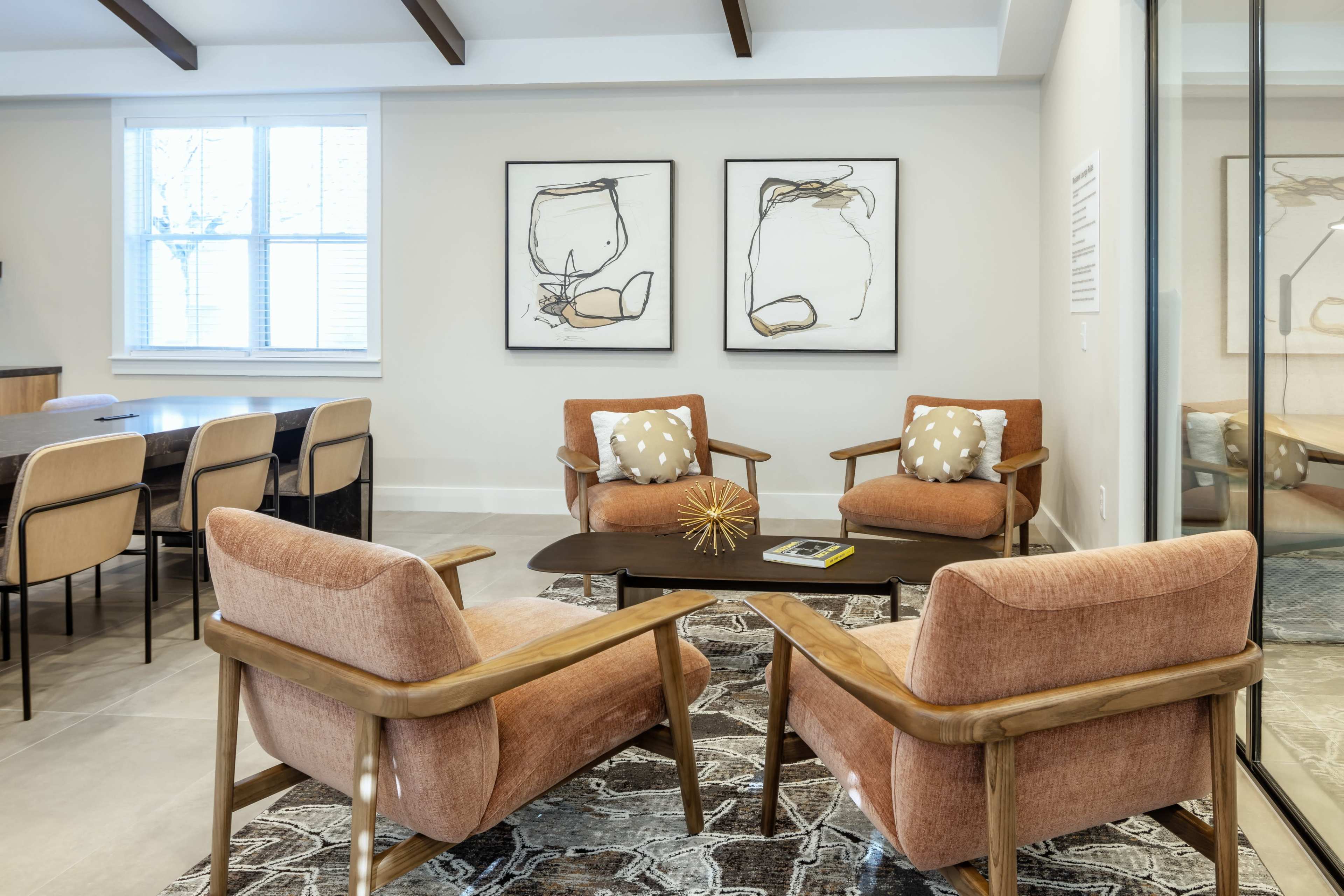 Two orange armchairs with pillows are placed around a dark wooden table in a modern living space, accompanied by abstract art on the walls and a dining area in the background.