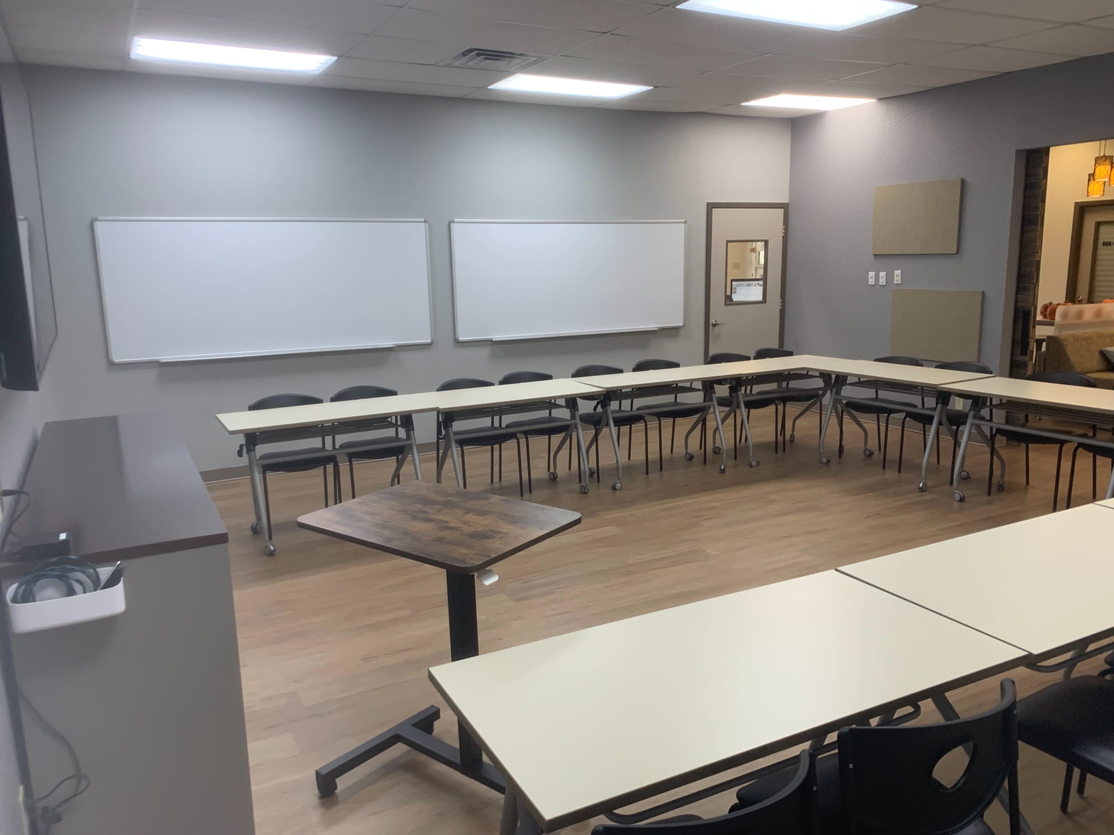 A classroom with a circular arrangement of tables and chairs, two whiteboards on the front wall, and a flat-screen monitor.
