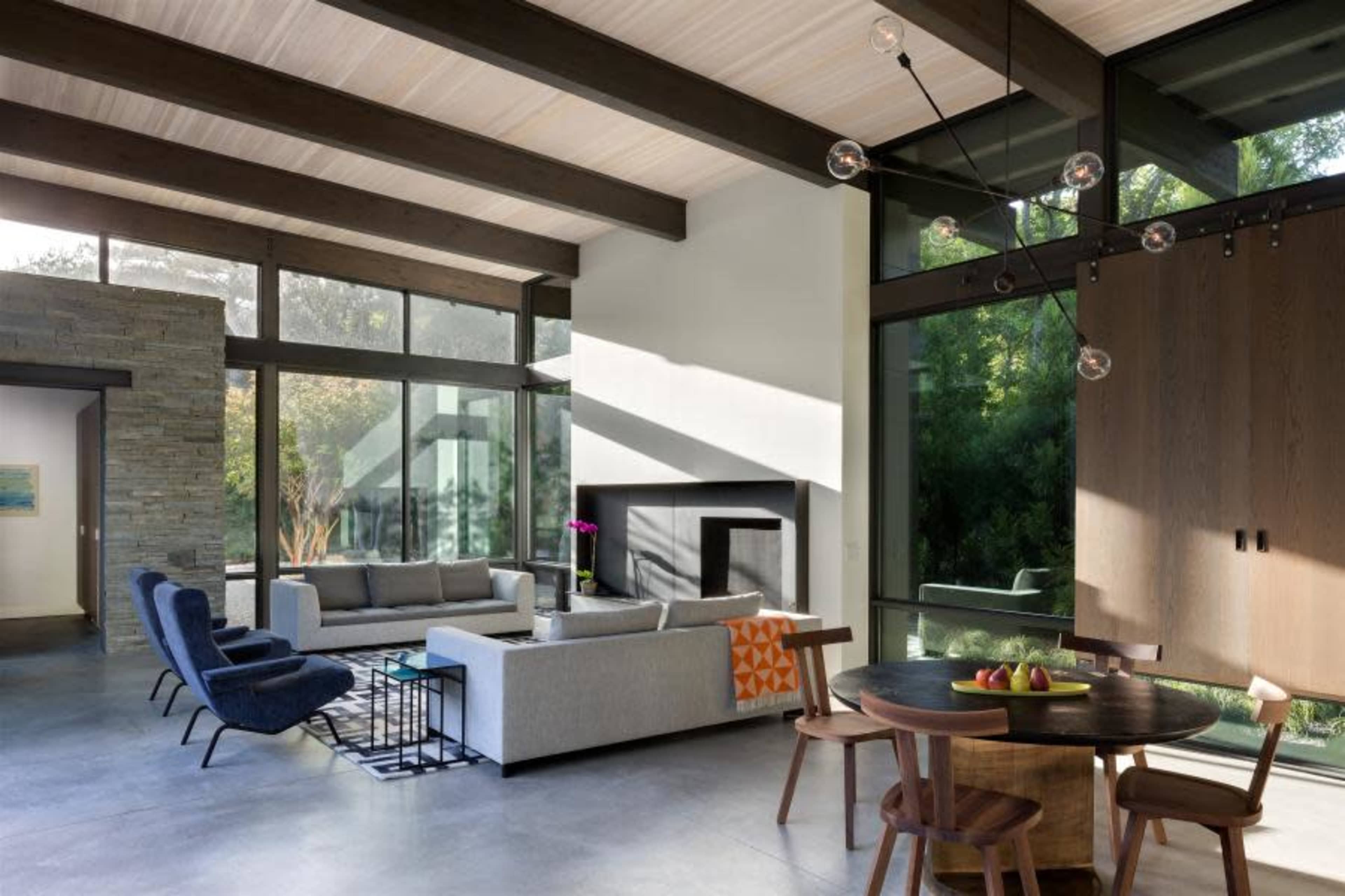 A modern living room features large windows, a stone accent wall, a grey sofa, blue chairs, a circular dining table, and a minimalist design.