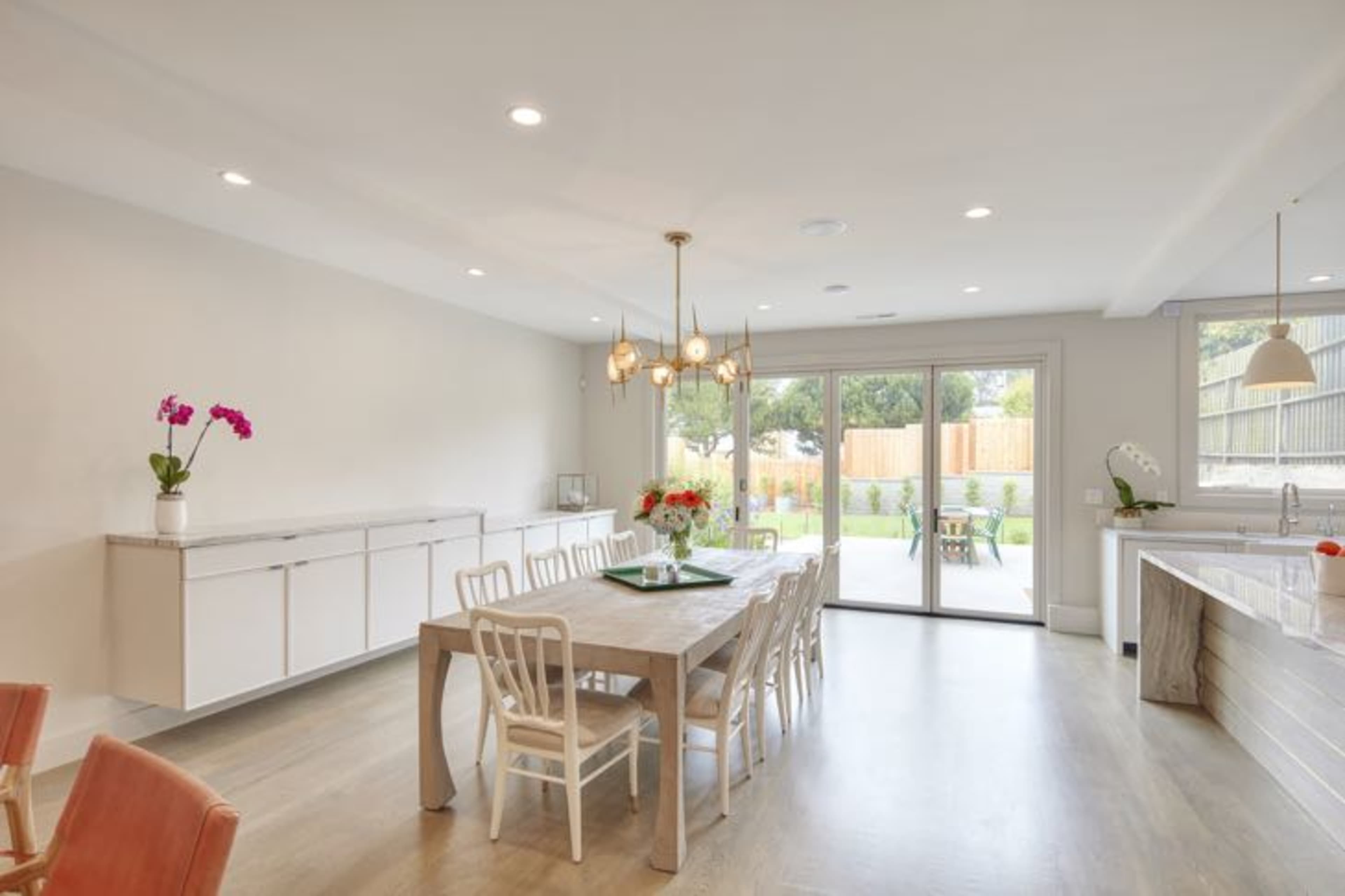 A modern dining area features a long table surrounded by chairs, with large sliding glass doors that open to a landscaped backyard.