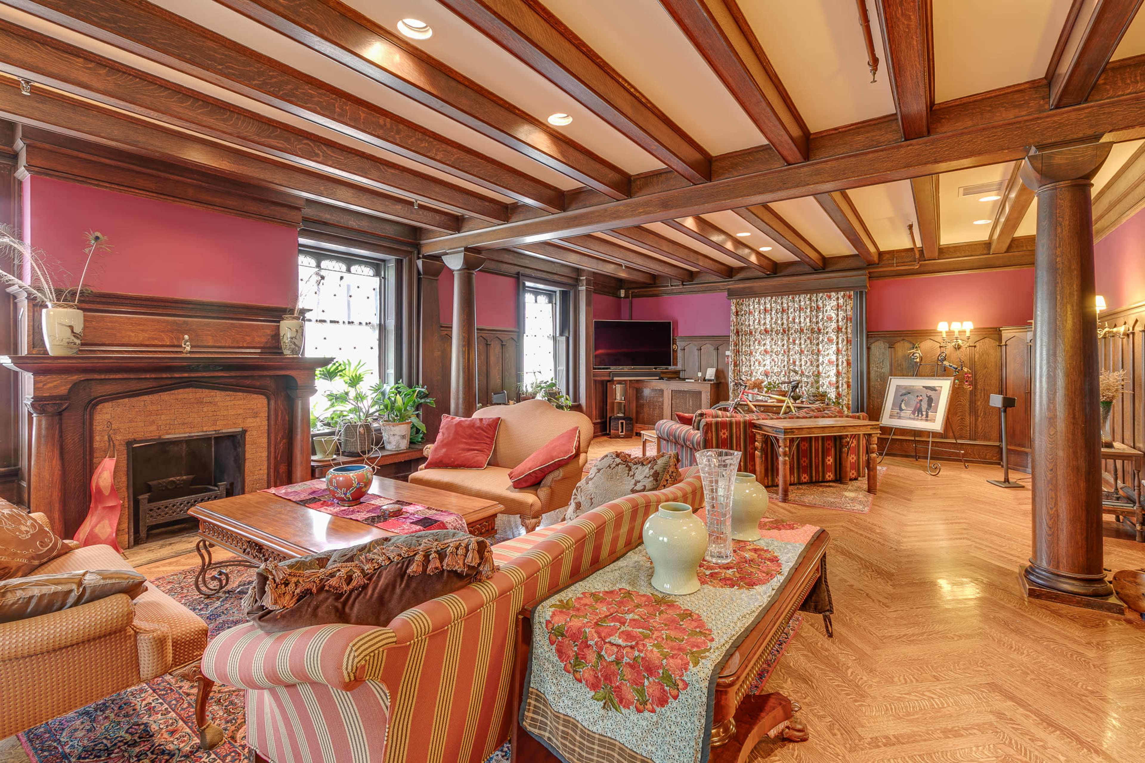 The image shows a spacious living room with wooden beam ceilings, a central fireplace, and a combination of seating arrangements around a coffee table.