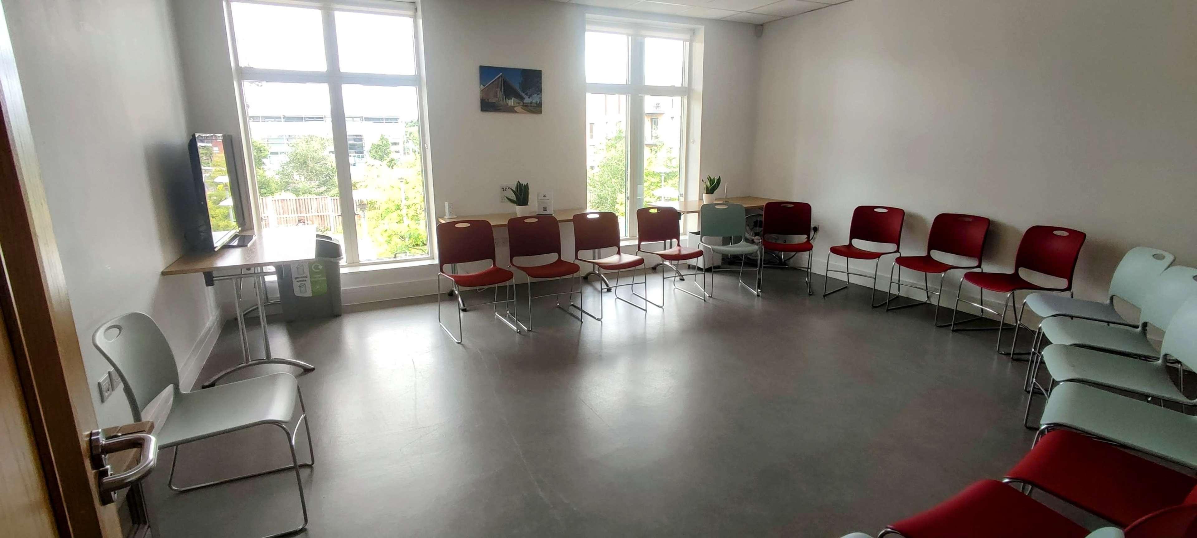 A meeting room features a circular arrangement of red and light blue chairs around a small table, with large windows allowing natural light to enter.