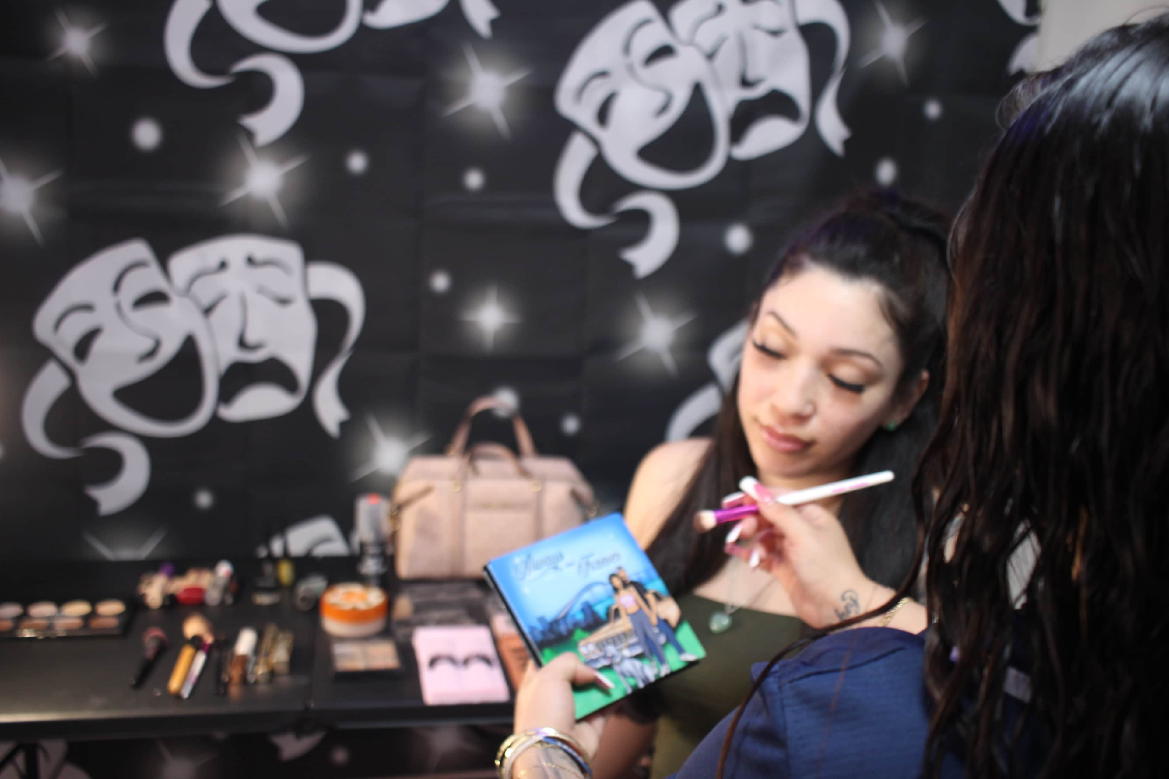 A makeup artist applies makeup to a client in front of a backdrop featuring theatrical masks, with various makeup products displayed on a table.