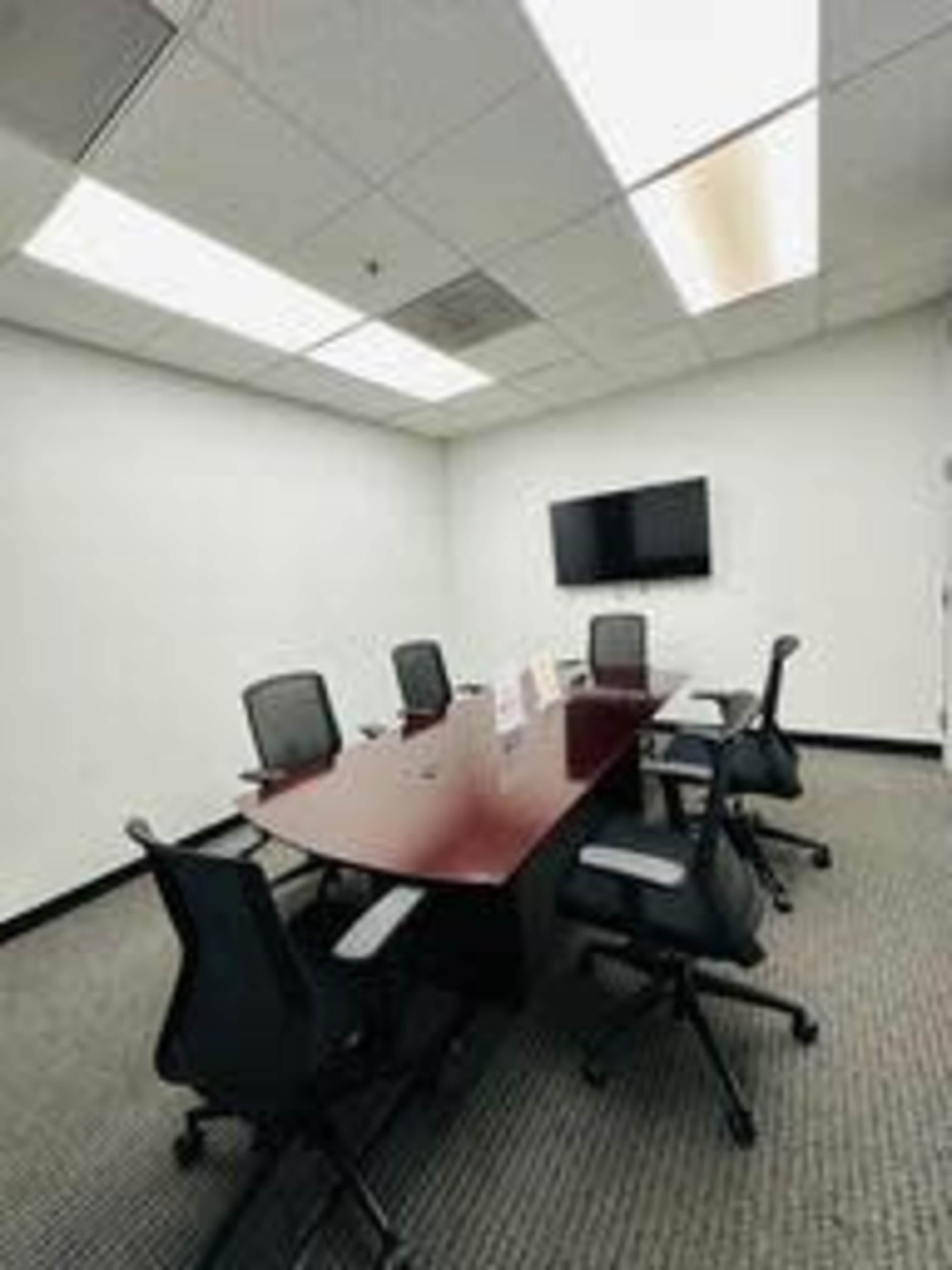 The image shows a conference room with a rectangular wooden table surrounded by six ergonomic chairs and a wall-mounted television.