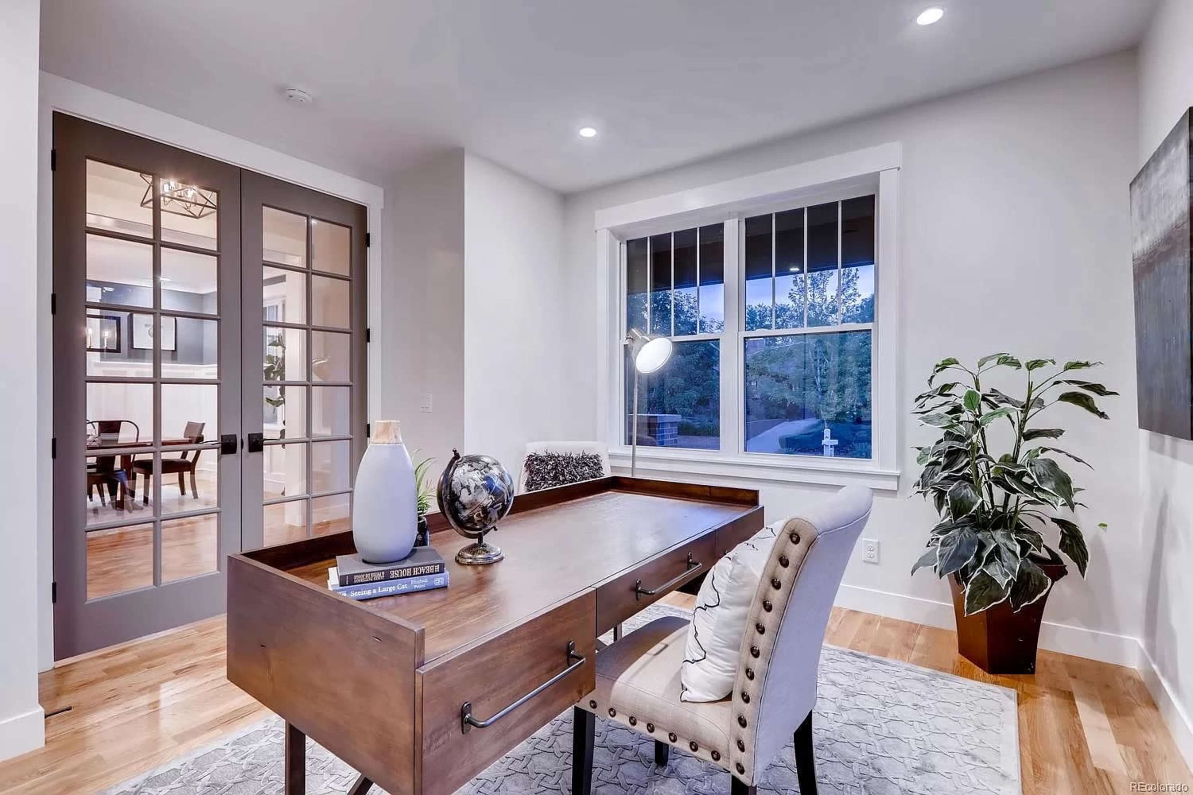 A modern home office features a wooden desk, a globe, a plant, and large windows overlooking a well-lit space.