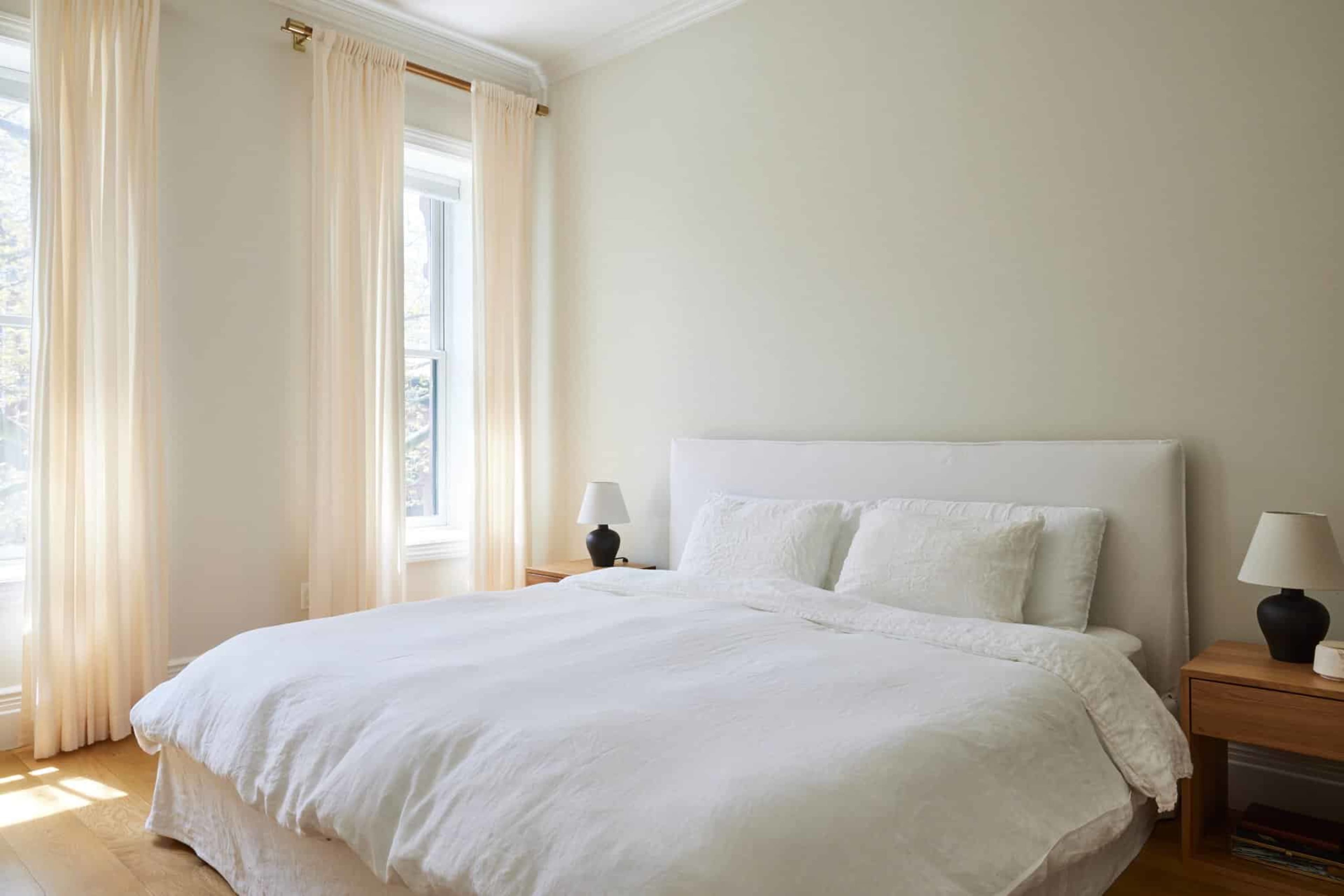 The image shows a bright, minimalist bedroom with a white bedspread, two small bedside lamps, and sheer curtains over the windows.