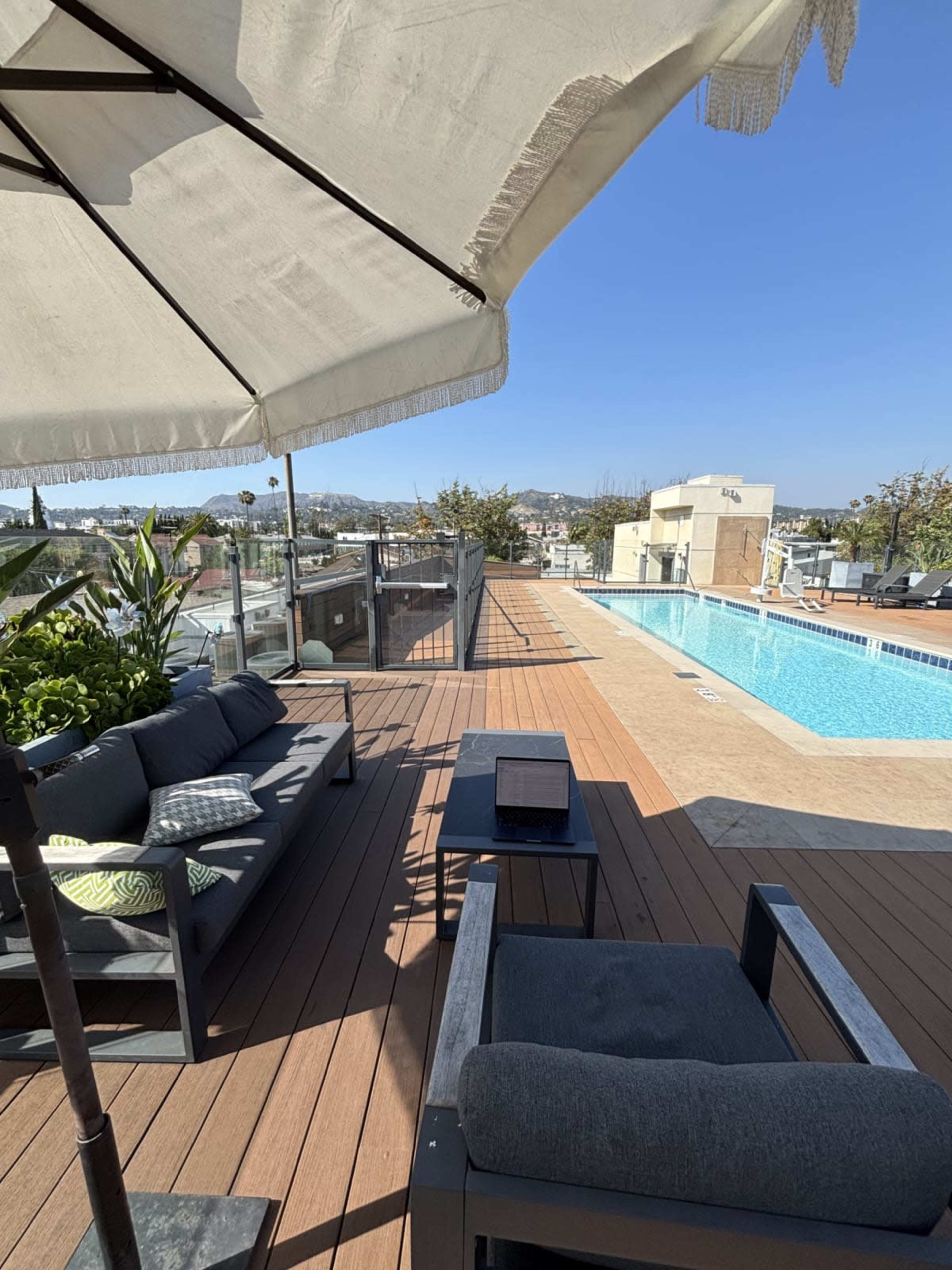A rooftop terrace features a swimming pool, modern outdoor furniture, and a view of a city skyline under a clear blue sky.