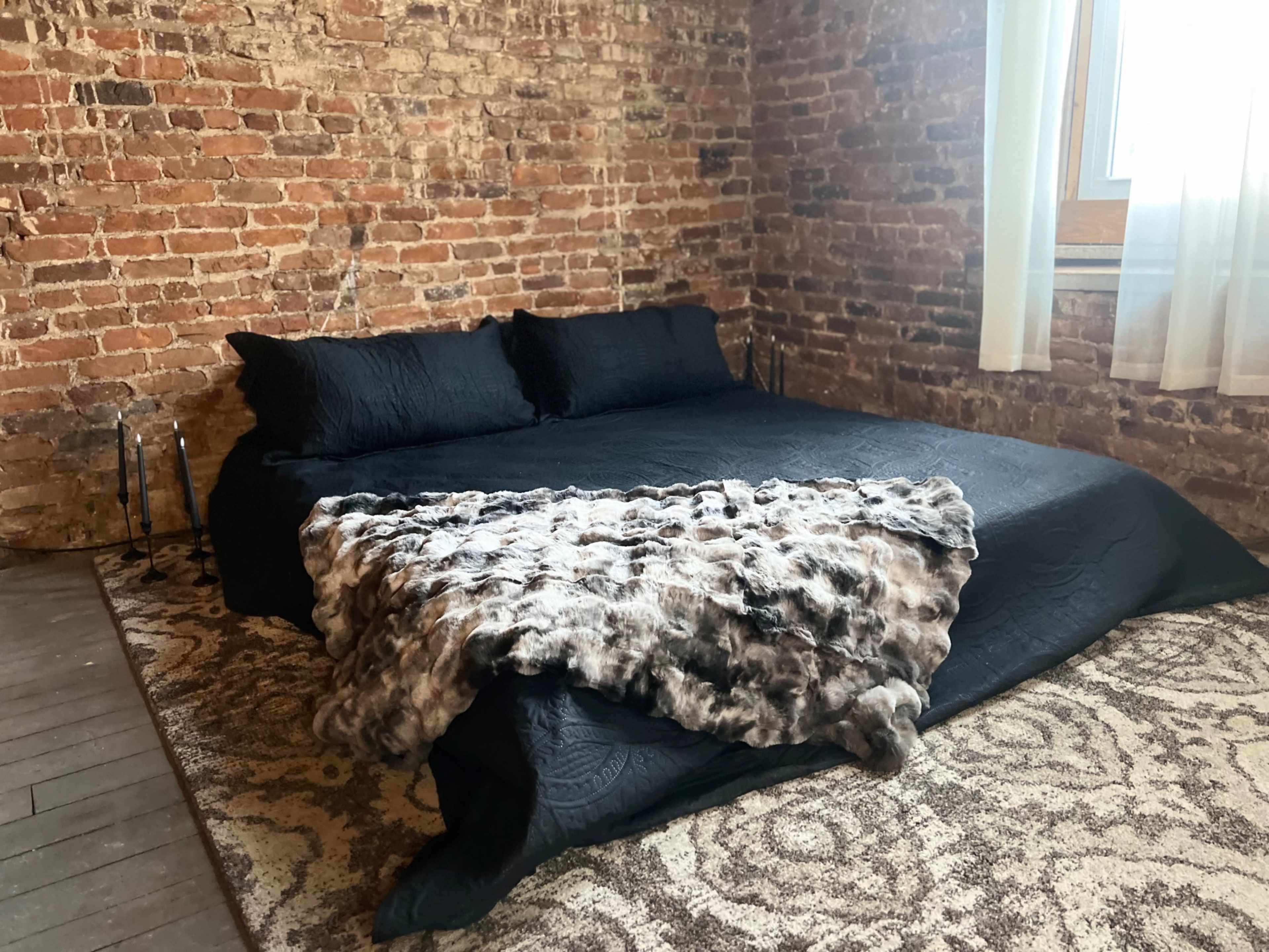 The image shows a bed with black bedding and a furry throw blanket in a room with exposed brick walls and a window.