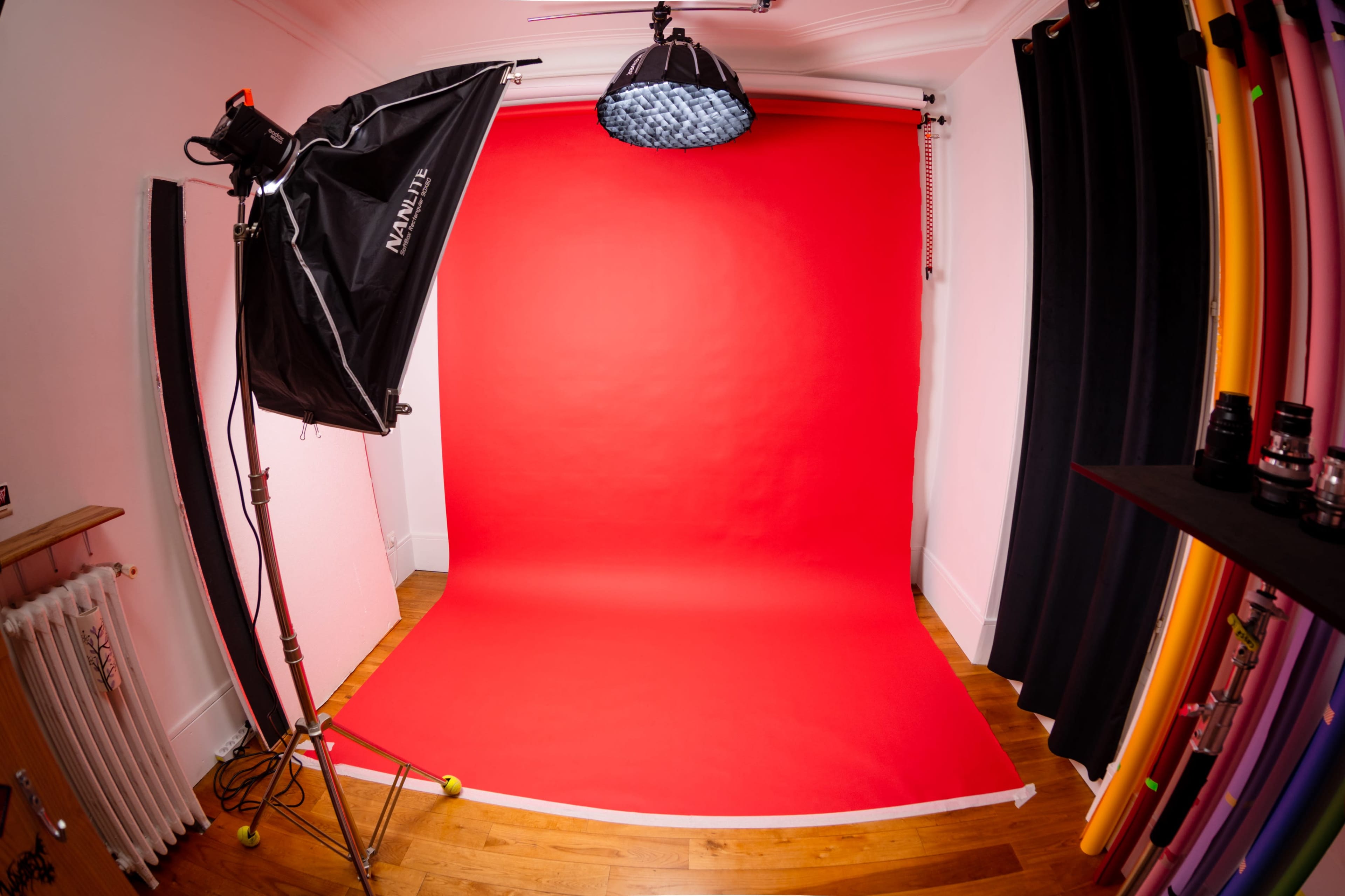 The image shows a photography studio setup with a red backdrop, lighting equipment, and various colored backdrops displayed on the side.