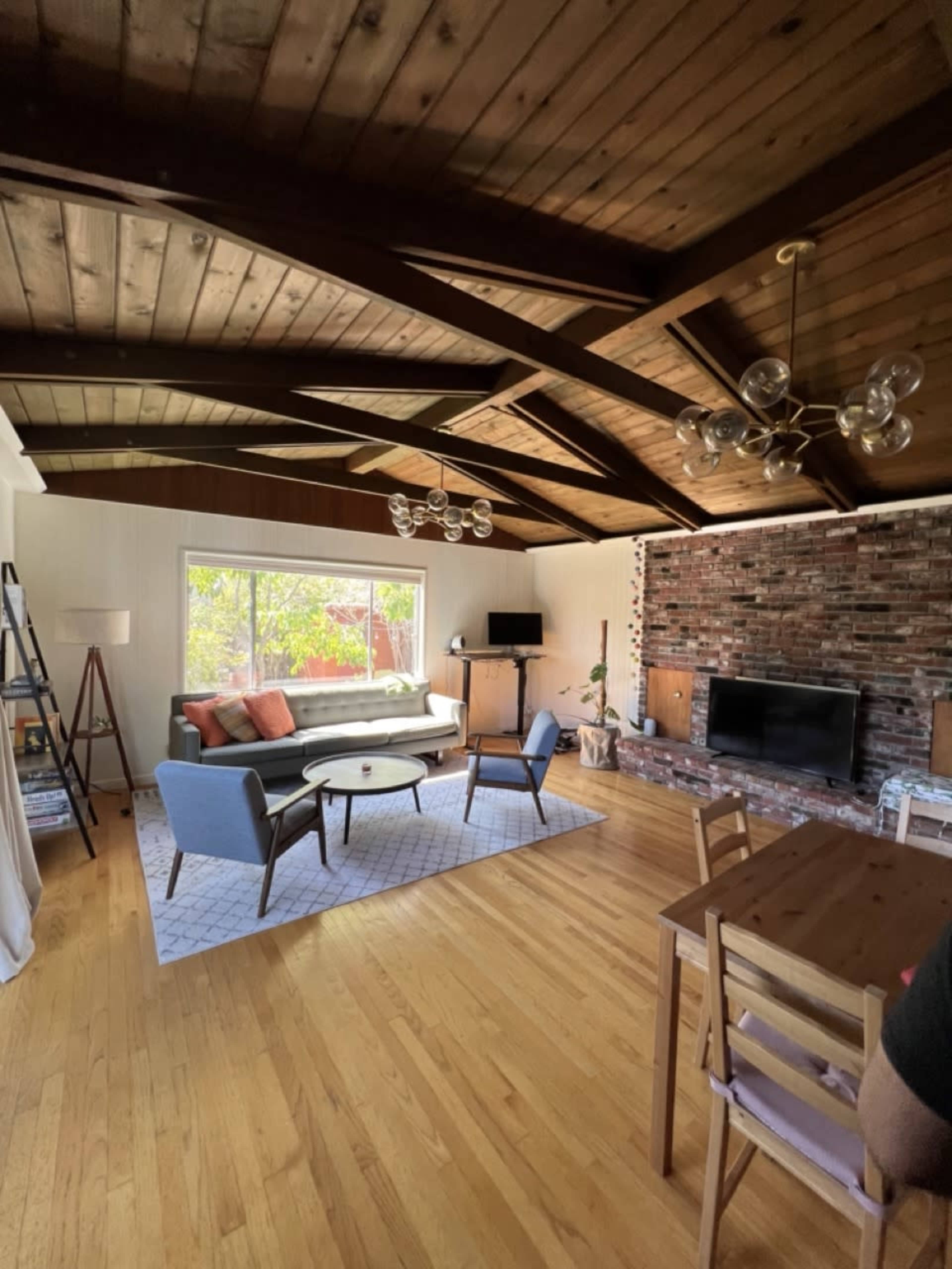 A spacious living room with a vaulted wooden ceiling, a large window, a brick accent wall, and a combination of modern and mid-century furniture.