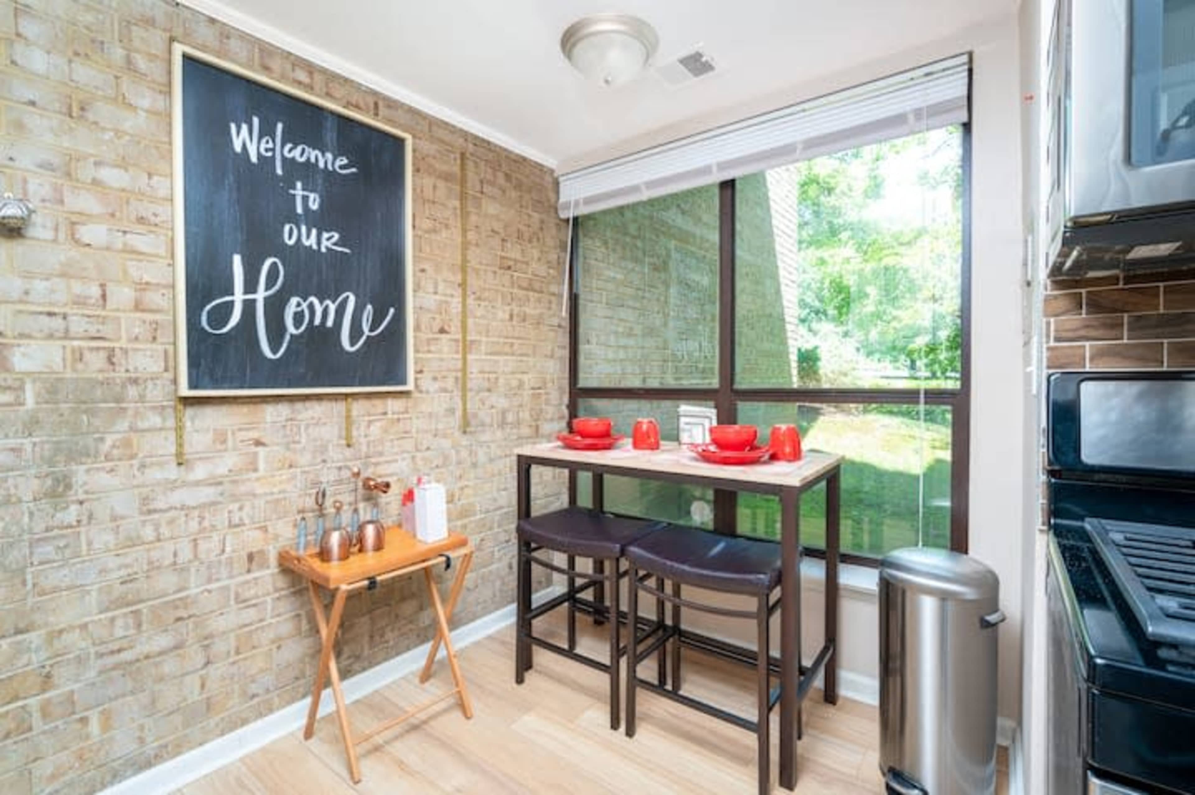 A small dining area with a table set for two, a chalkboard welcoming visitors, and a window that looks out onto a green area.