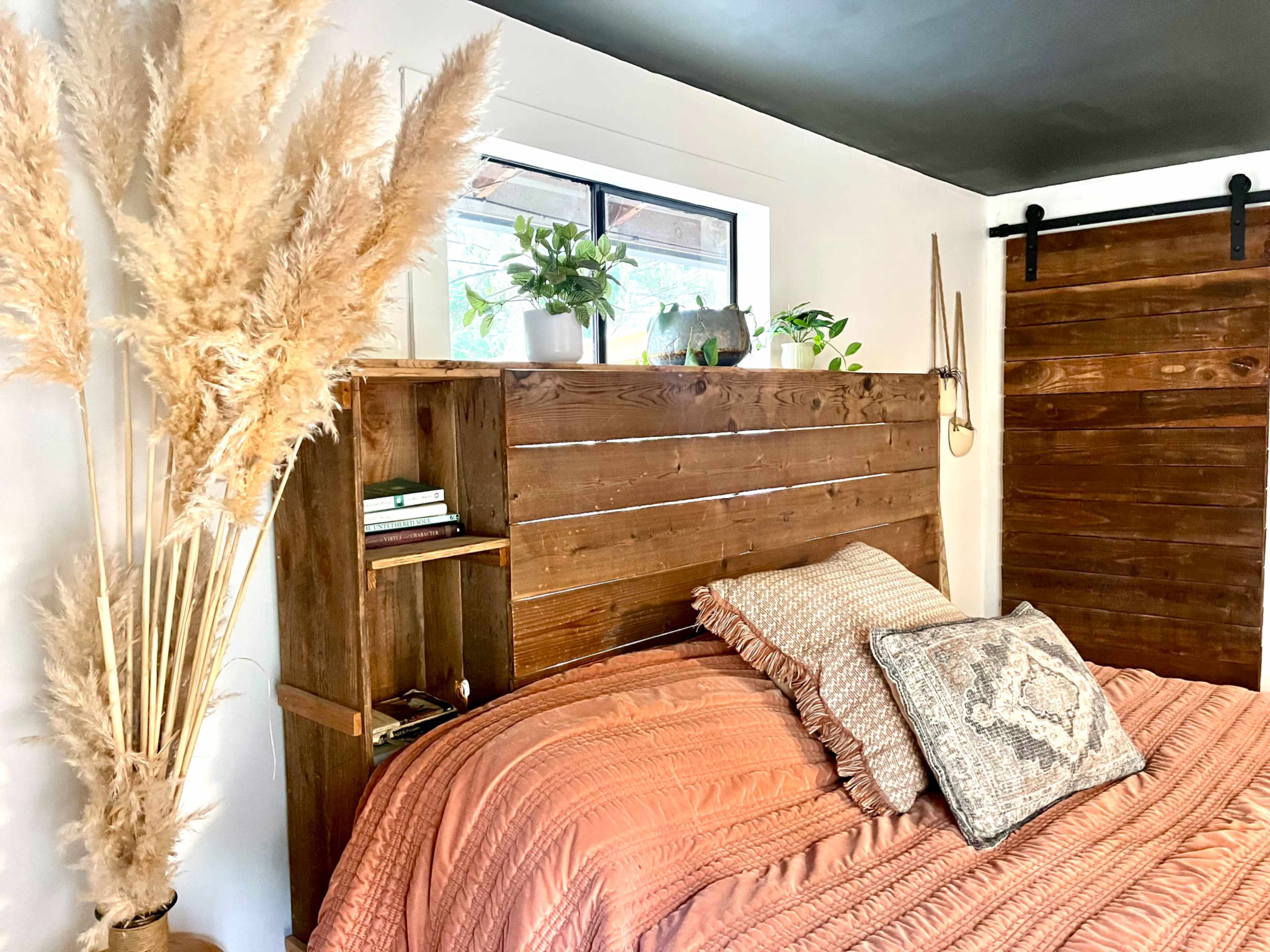The image shows a cozy bedroom featuring a wooden slatted headboard, an orange bedspread, and decorative plants on shelves beside the bed.