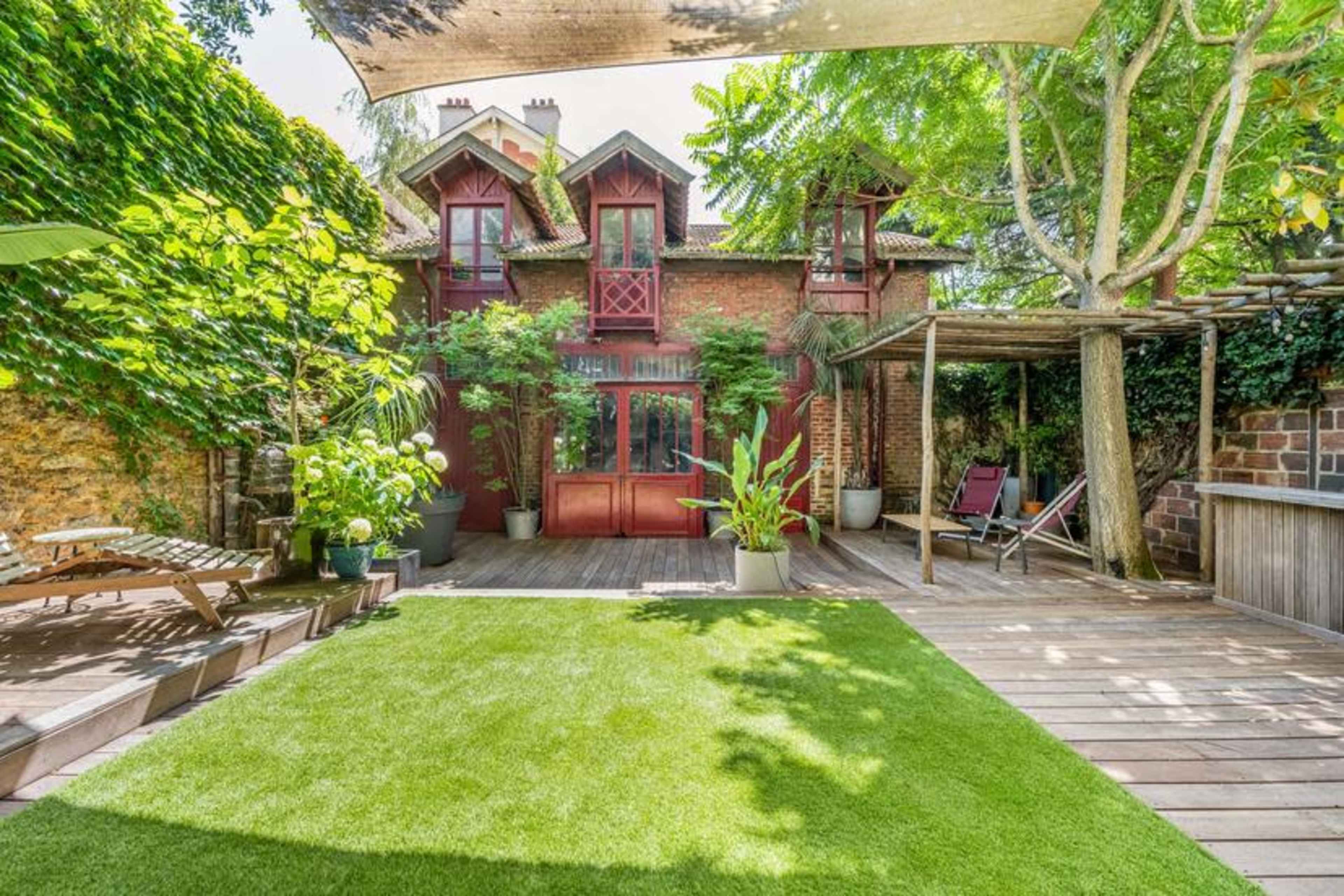 A backyard featuring a lush green lawn, wooden decking, and a brick house with red trim and multiple windows.