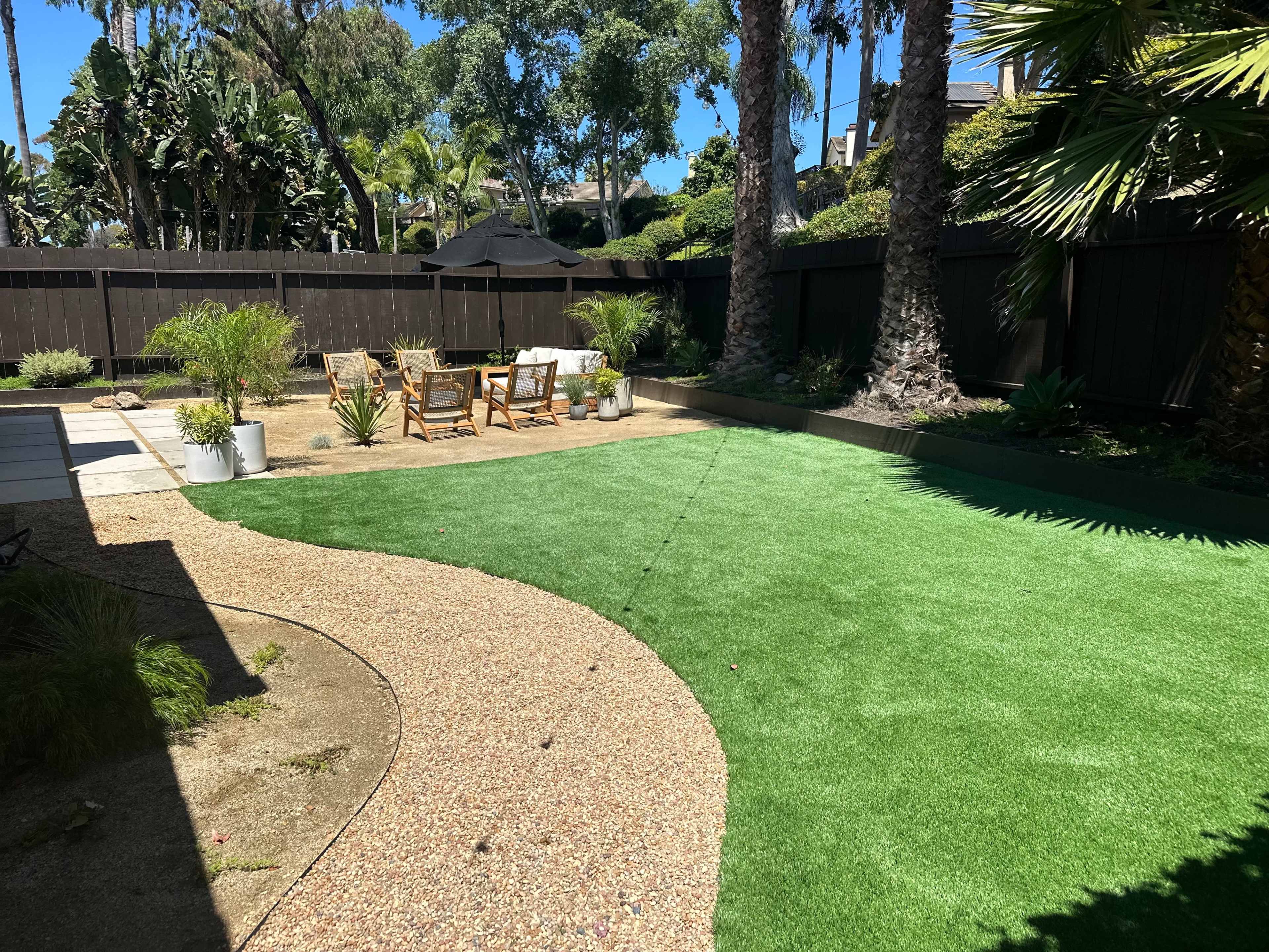 A landscaped backyard featuring a pathway made of gravel, artificial turf, potted plants, and seating arrangements under palm trees.