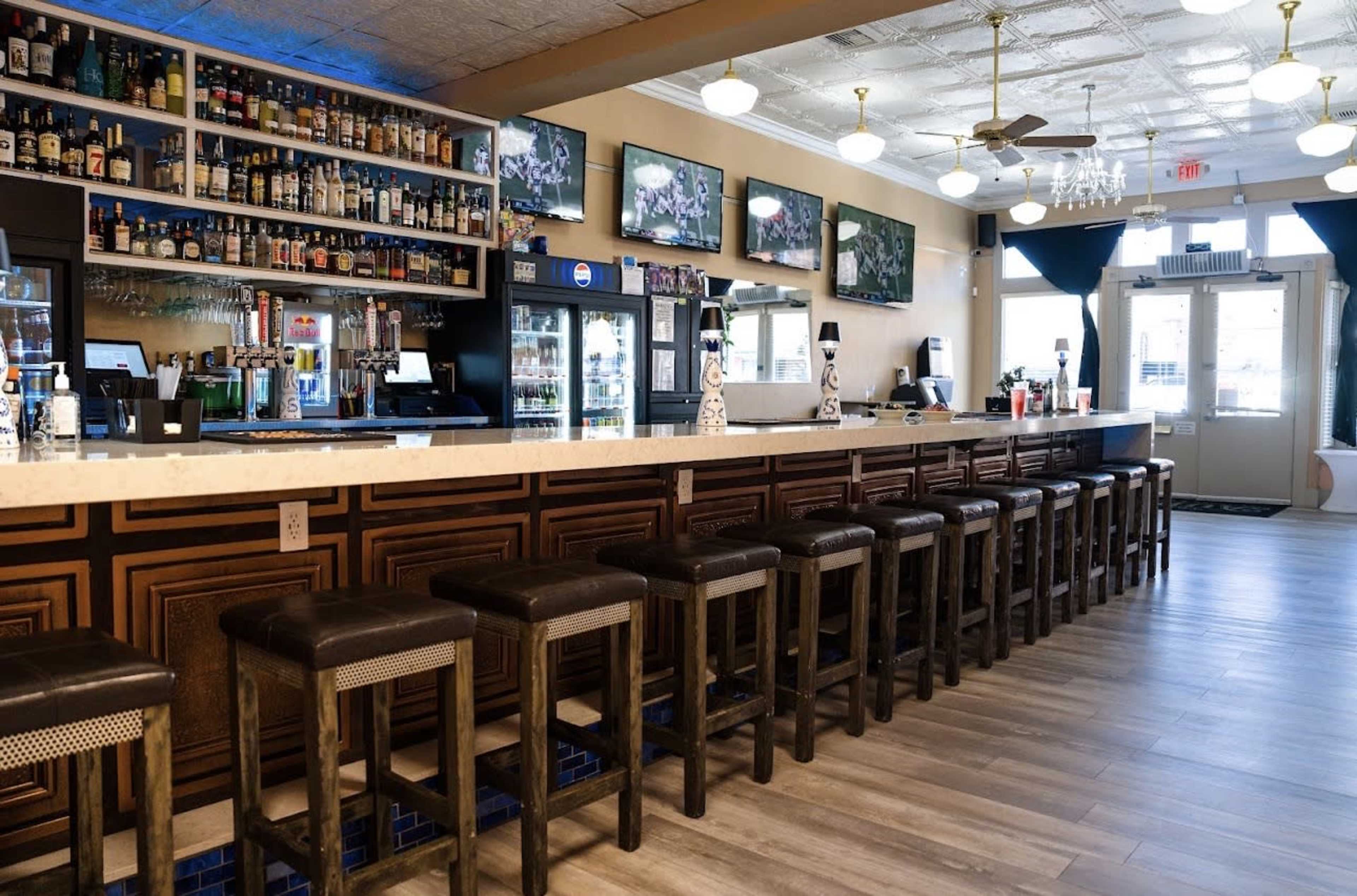 The image shows a modern bar area featuring a long counter with high stools, shelves displaying various bottles, and multiple screens mounted on the walls.