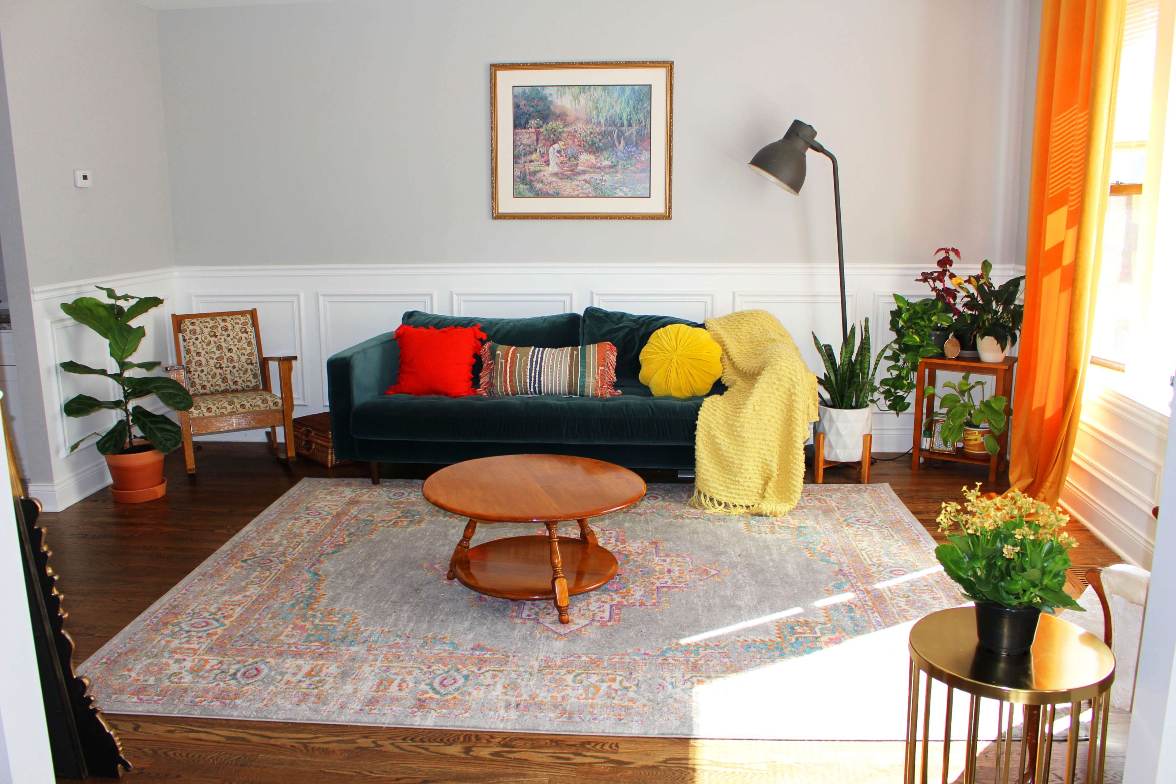 The living room features a green sofa with colorful cushions, a round wooden coffee table, a patterned area rug, and various plants in decorative pots.