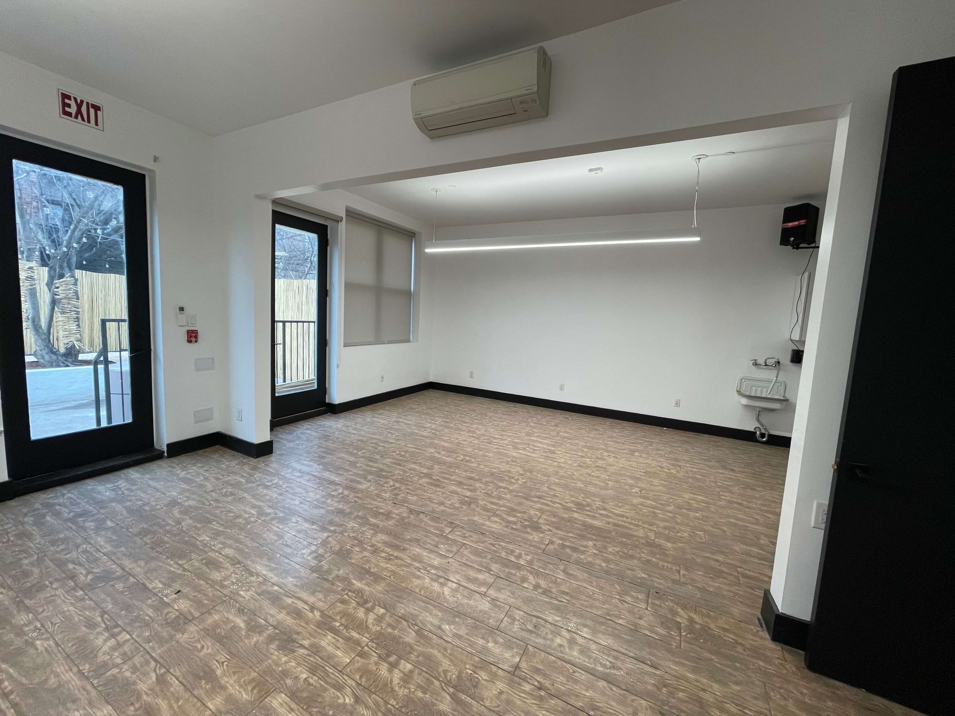 The image shows a vacant room with wooden flooring, large windows, and minimal furniture, featuring an exit sign and an air conditioning unit.