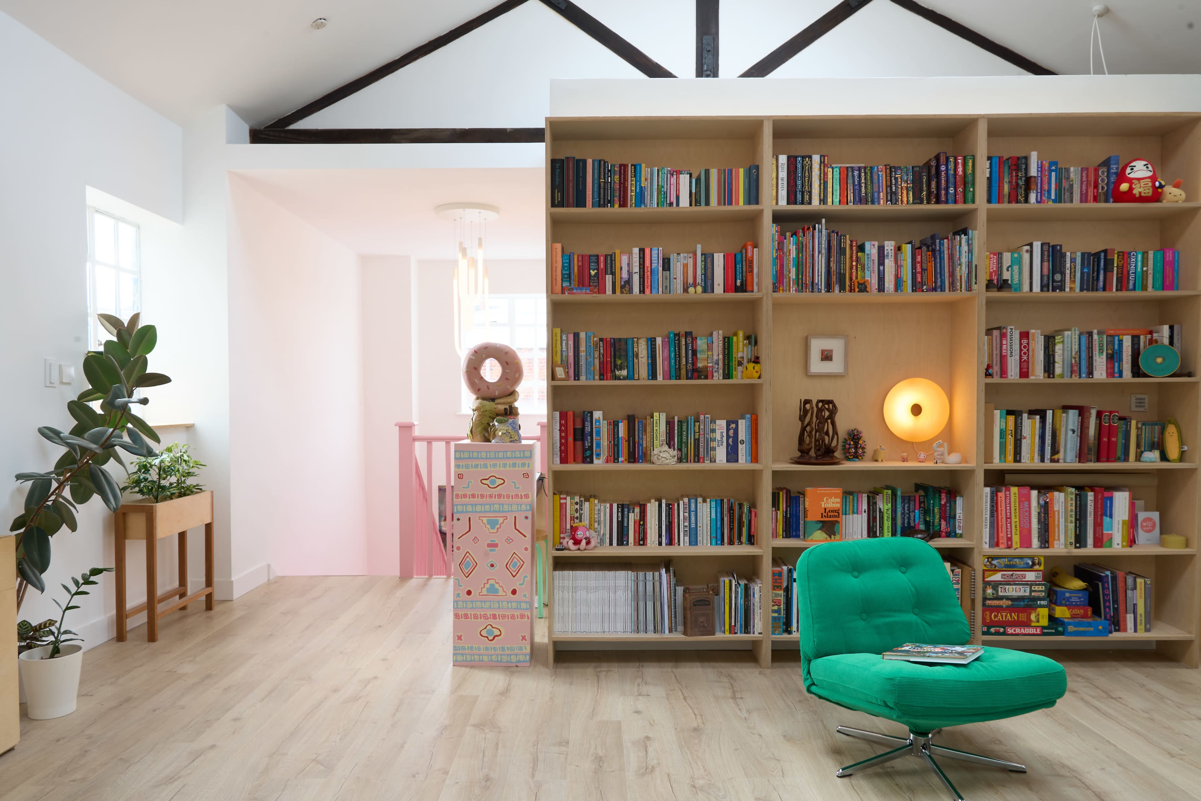 The image shows a bright, modern interior featuring a well-organized bookshelf filled with colorful books, a green lounge chair, and a small table with a plant.